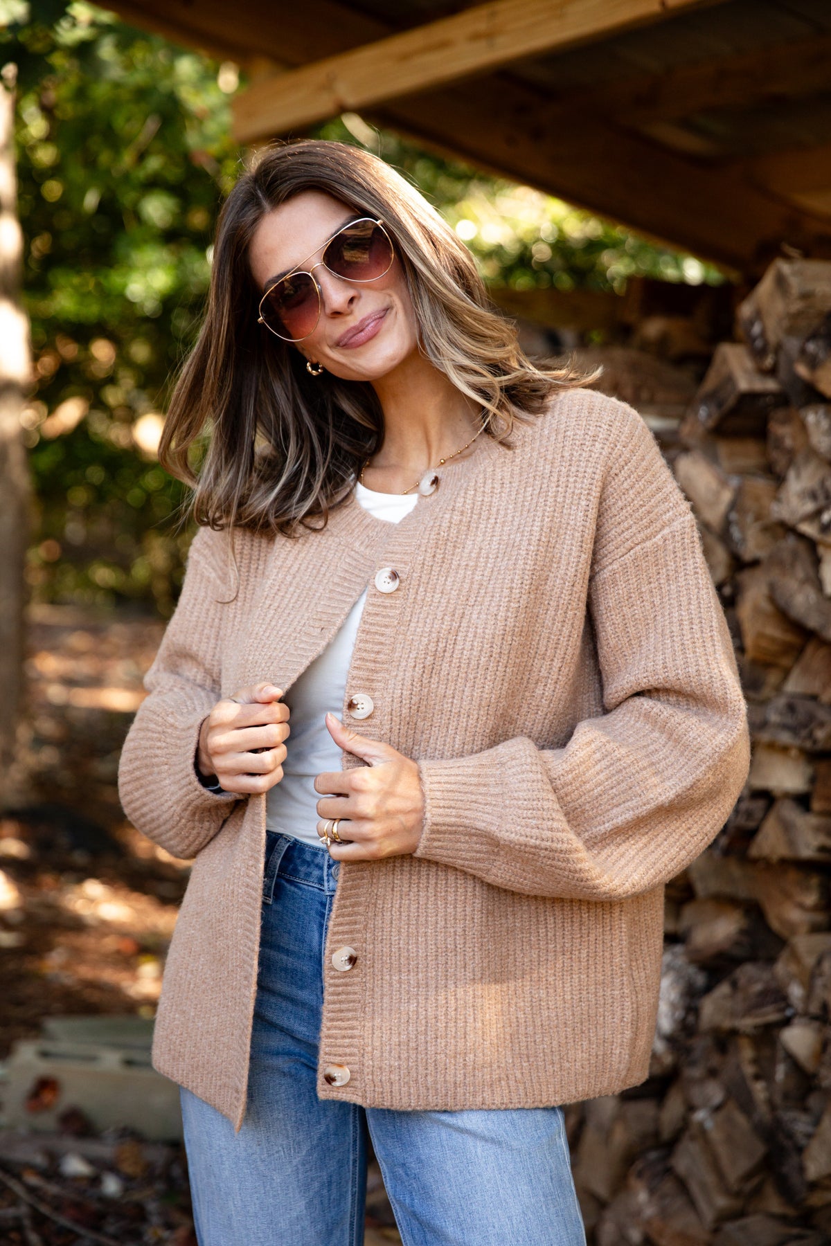 Woman wearing a beige cardigan and sunglasses outdoors near stacked firewood.