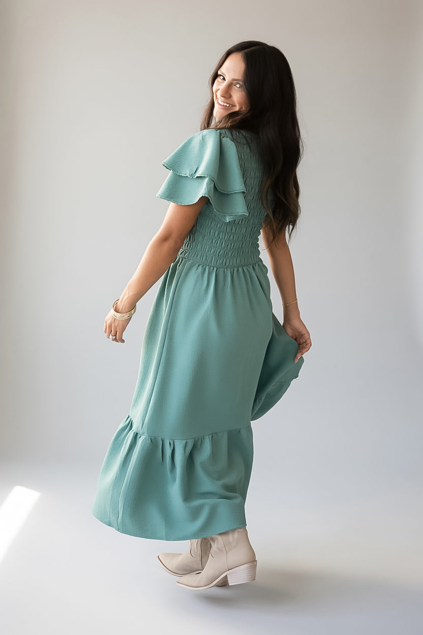 A woman wearing a sage green tiered maxi dress with ruffled sleeves and a smocked bodice, paired with beige ankle boots, photographed against a light gray studio background.