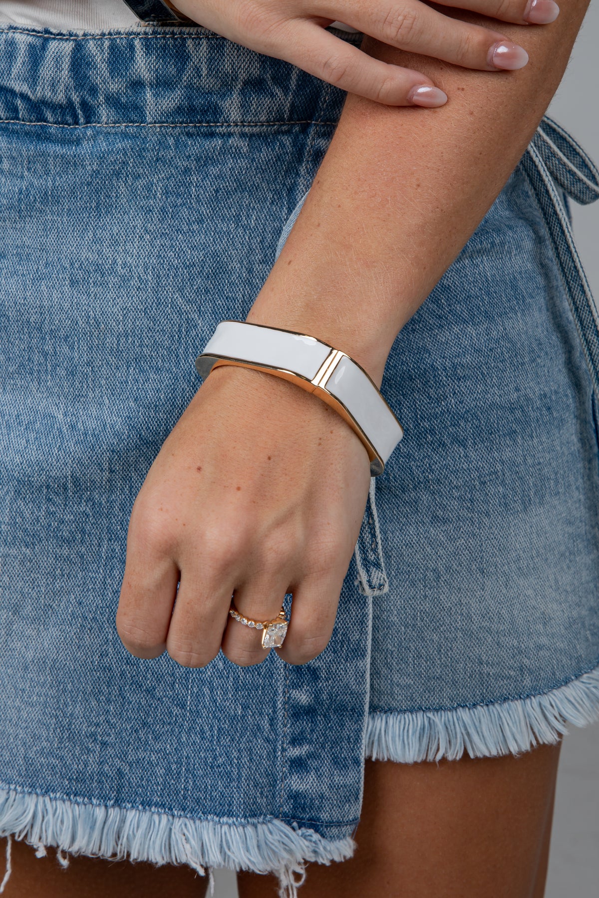 A close-up of a person wearing a wide white cuff bracelet with a gold-toned metal band and a gold accent bar, paired with a denim skirt.
