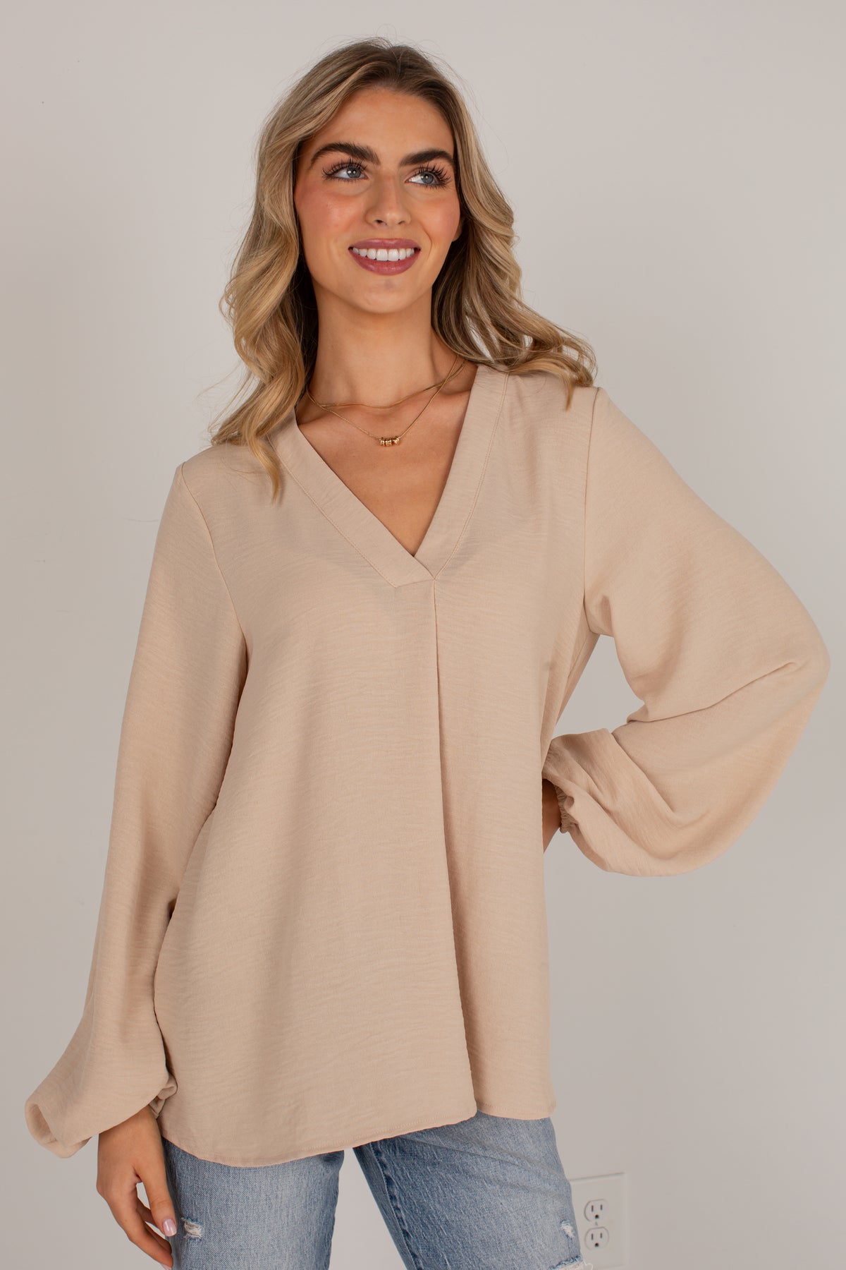 A woman wearing a light beige long-sleeve blouse with a V-neckline and balloon sleeves, paired with blue jeans, standing against a plain light gray background.