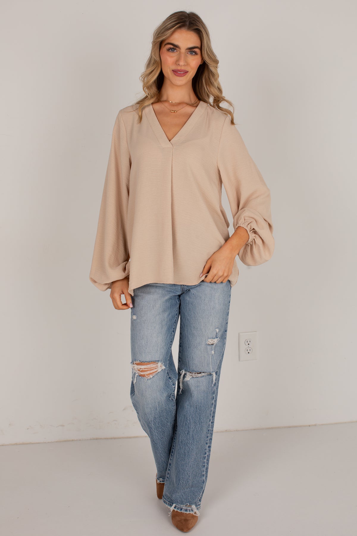 A woman modeling a beige long-sleeve blouse with a V-neck and balloon sleeves, paired with distressed blue jeans and brown shoes, standing against a plain light gray wall.