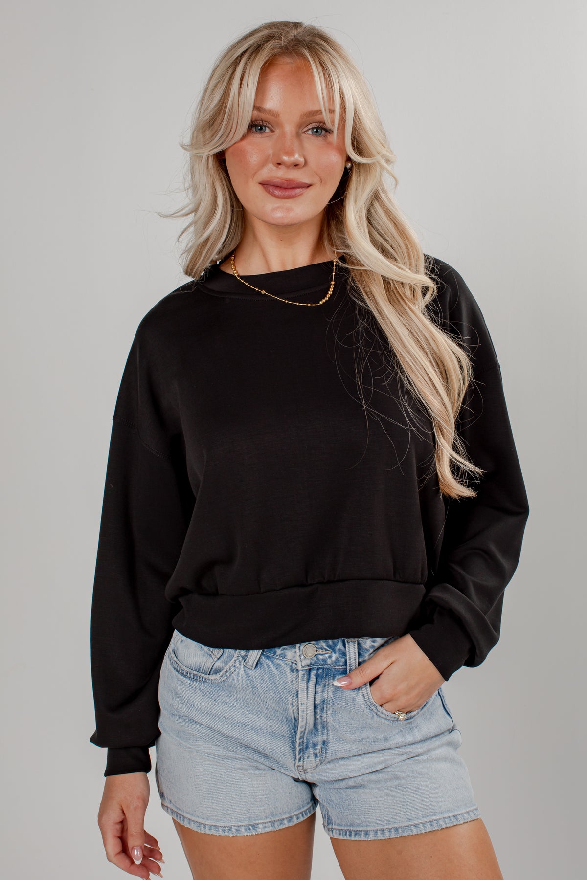 A blonde woman wearing a black long-sleeve crewneck sweatshirt and light-wash denim shorts, standing against a neutral gray studio background.