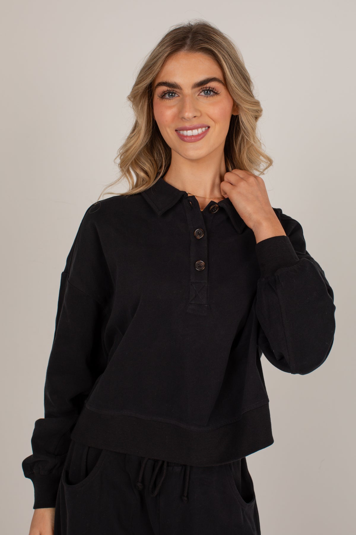 A woman wearing a black long-sleeve polo shirt with a buttoned collar, shown against a plain light gray studio background.