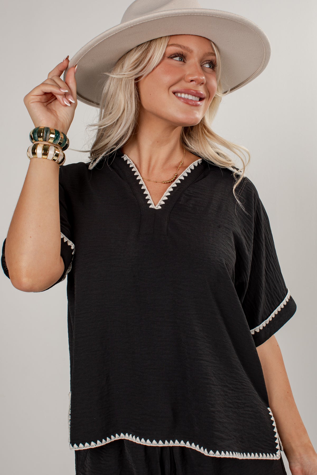 A woman wearing a black short-sleeve top with white geometric trim, a wide-brimmed beige hat, and gold bracelets, photographed against a light gray studio background.