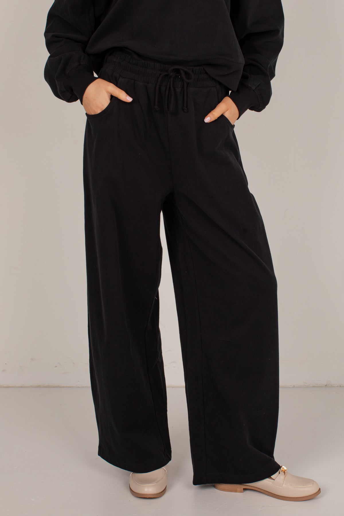 A person wearing loose black wide-leg drawstring pants with a black sweatshirt and beige loafers, standing against a plain light gray studio background.