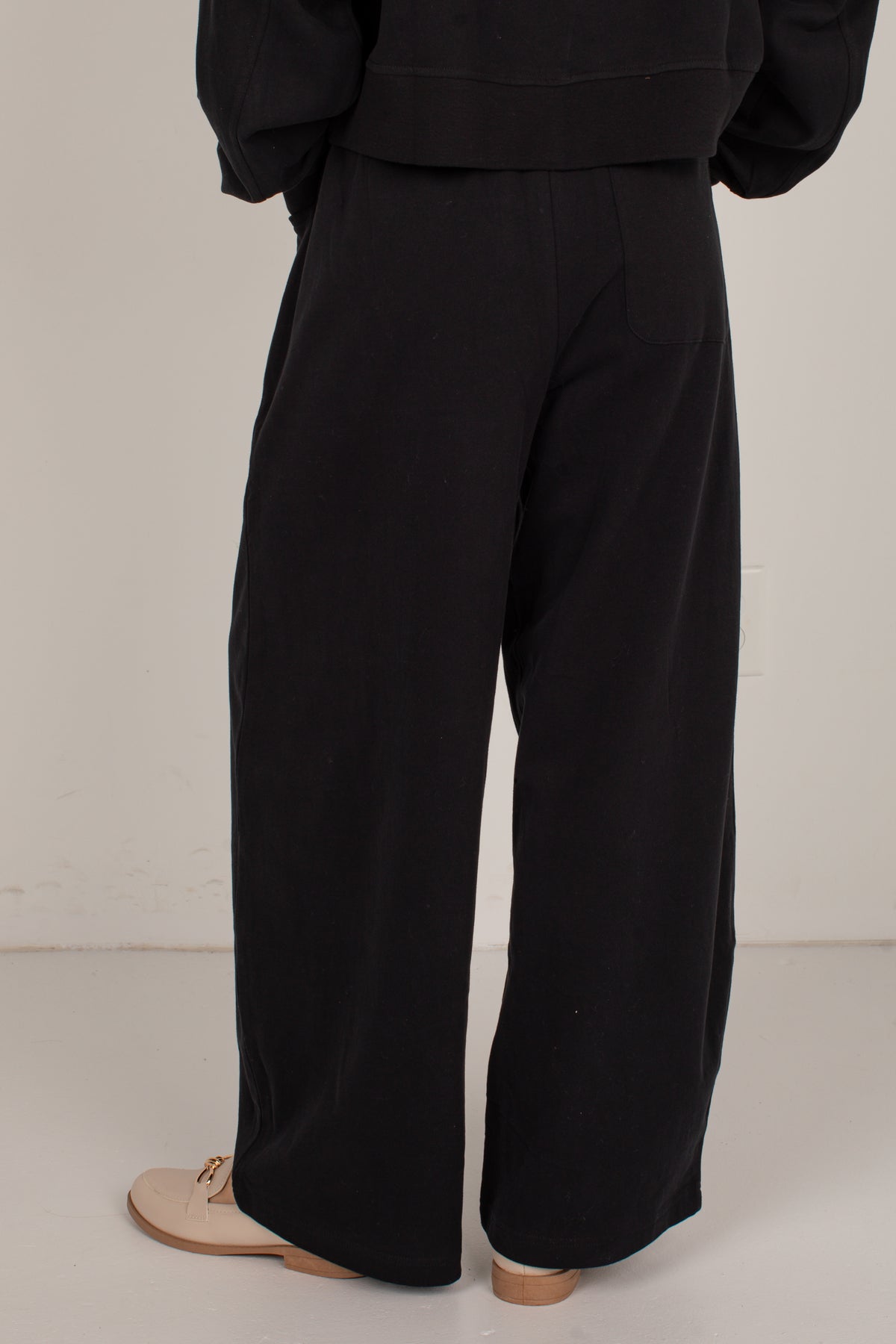 Black wide-leg trousers shown from the back on a model, paired with a black top and beige flat shoes, against a plain off-white studio background.