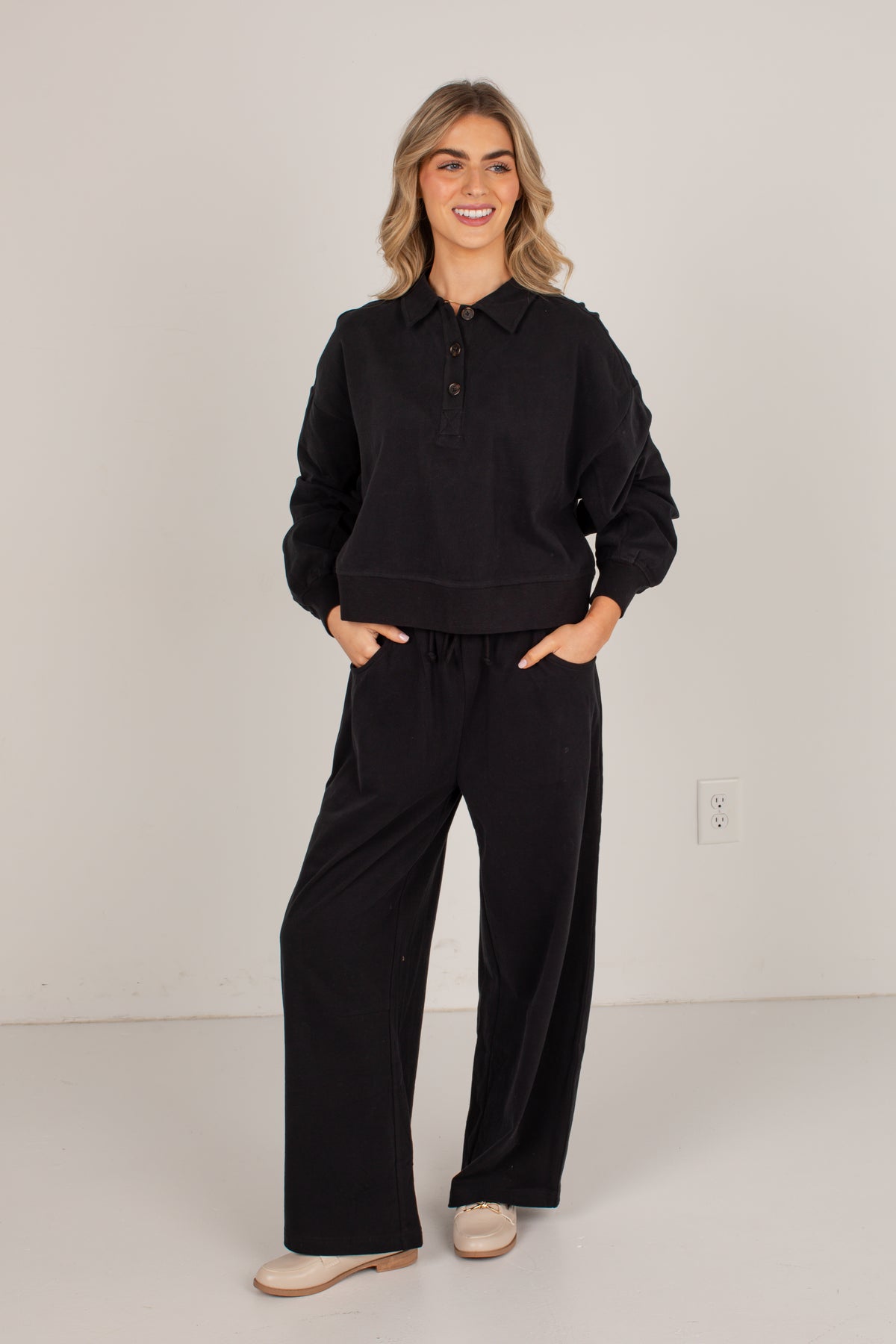 A woman modeling a black long-sleeve polo-style top and matching black wide-leg pants, standing against a plain light gray studio wall.