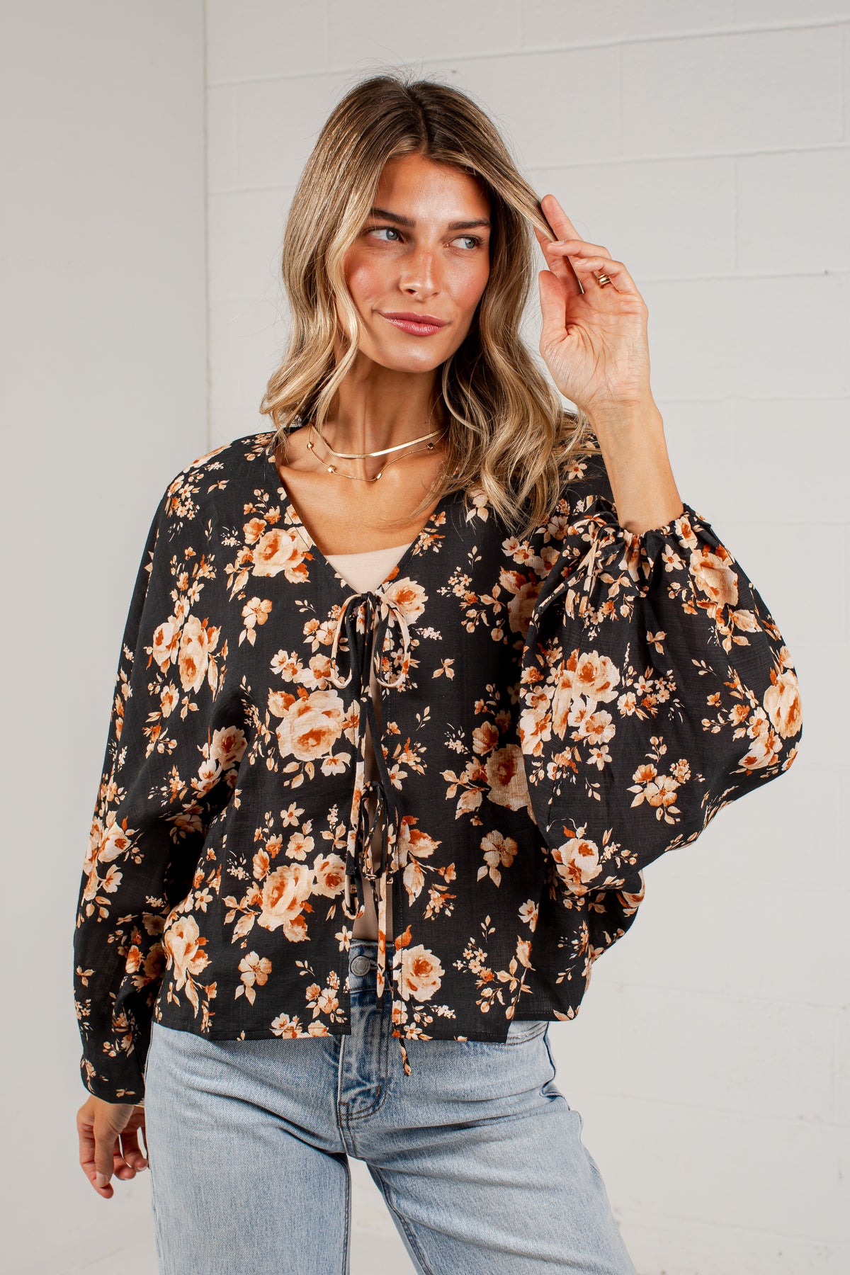A woman wearing a black long-sleeve blouse with a warm-toned orange and beige floral print, paired with light-wash blue jeans, standing against a white brick wall.
