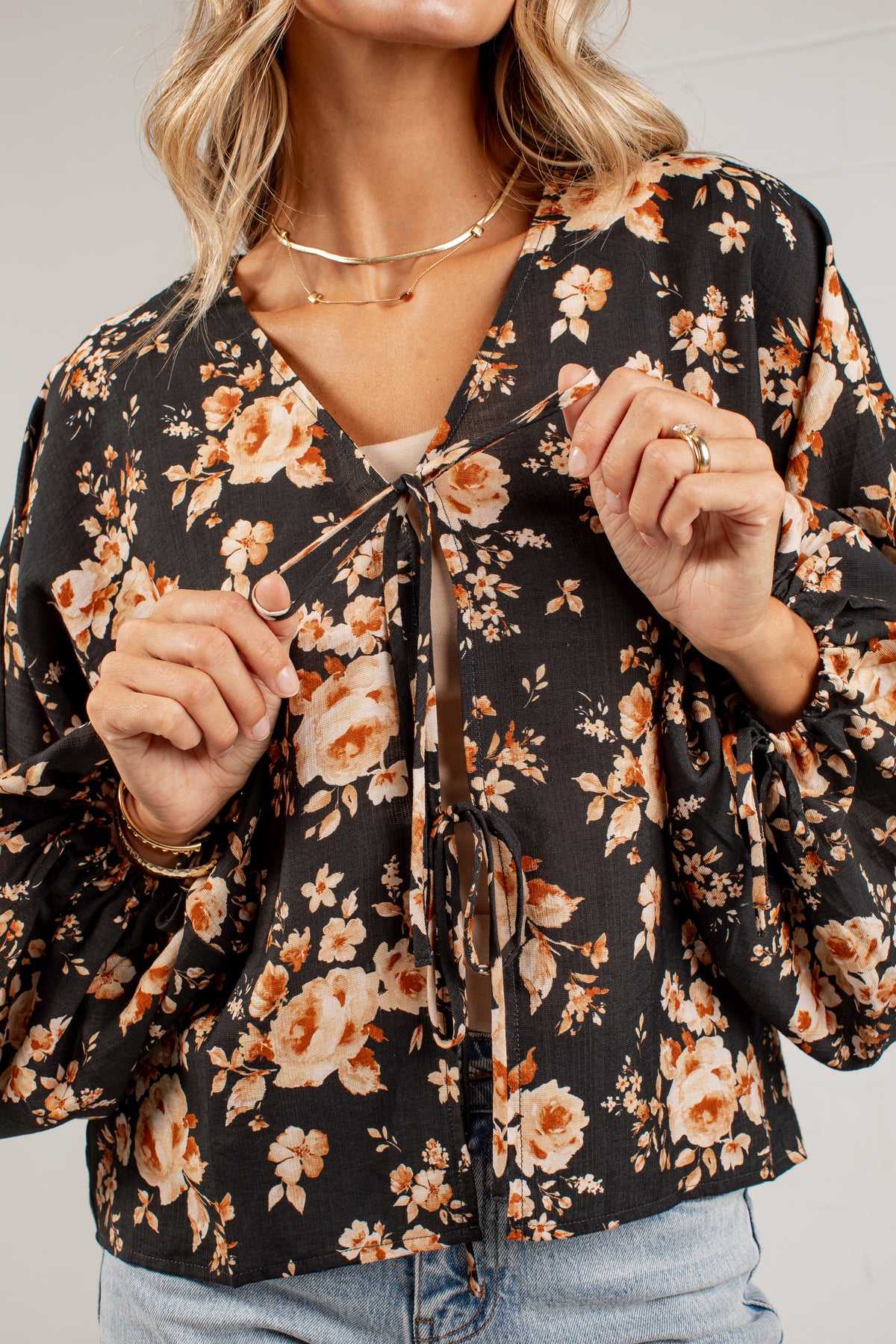 A woman wearing a black long-sleeve blouse with a warm-toned floral print in orange, beige, and cream, featuring a tie-front closure and voluminous sleeves.