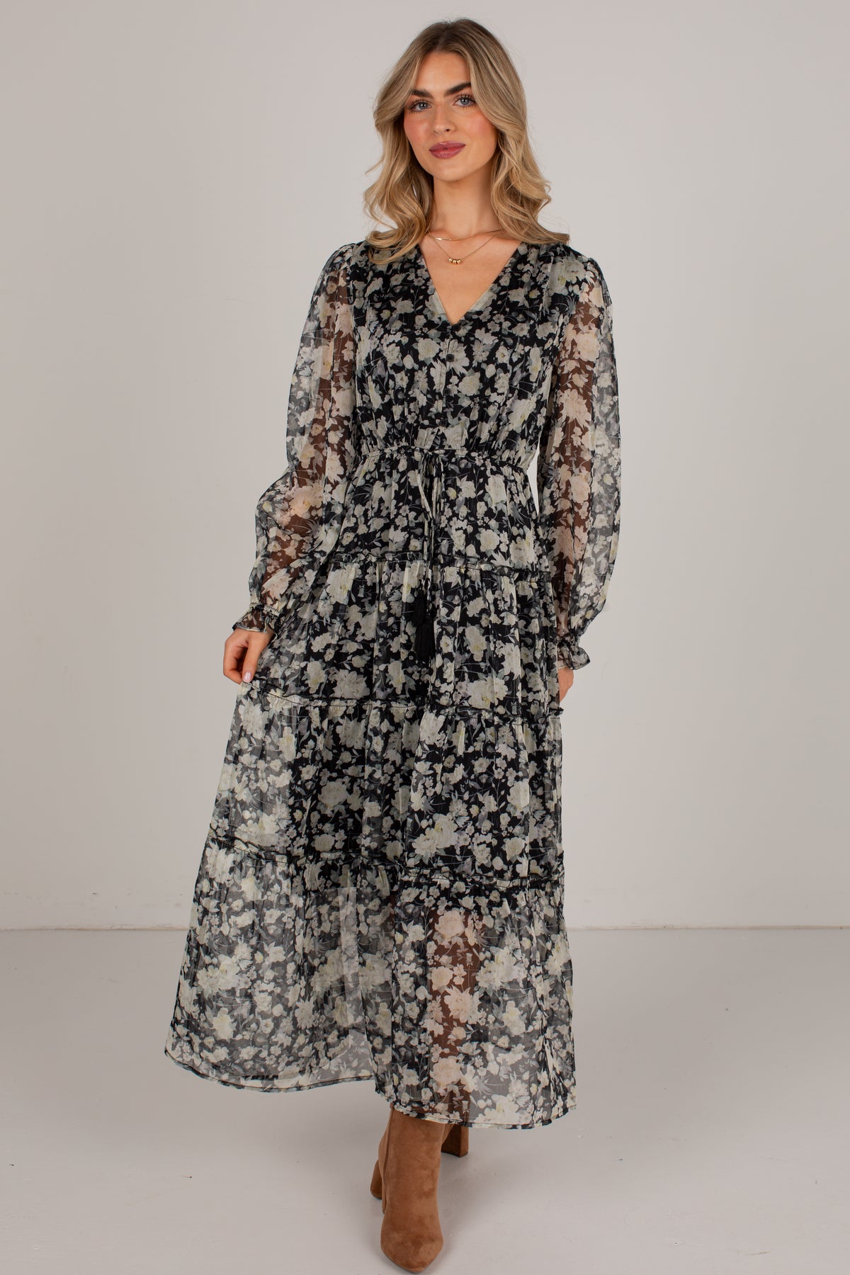 A woman modeling a long-sleeve midi dress with a black, gray, and beige floral pattern, standing against a plain light gray studio background.
