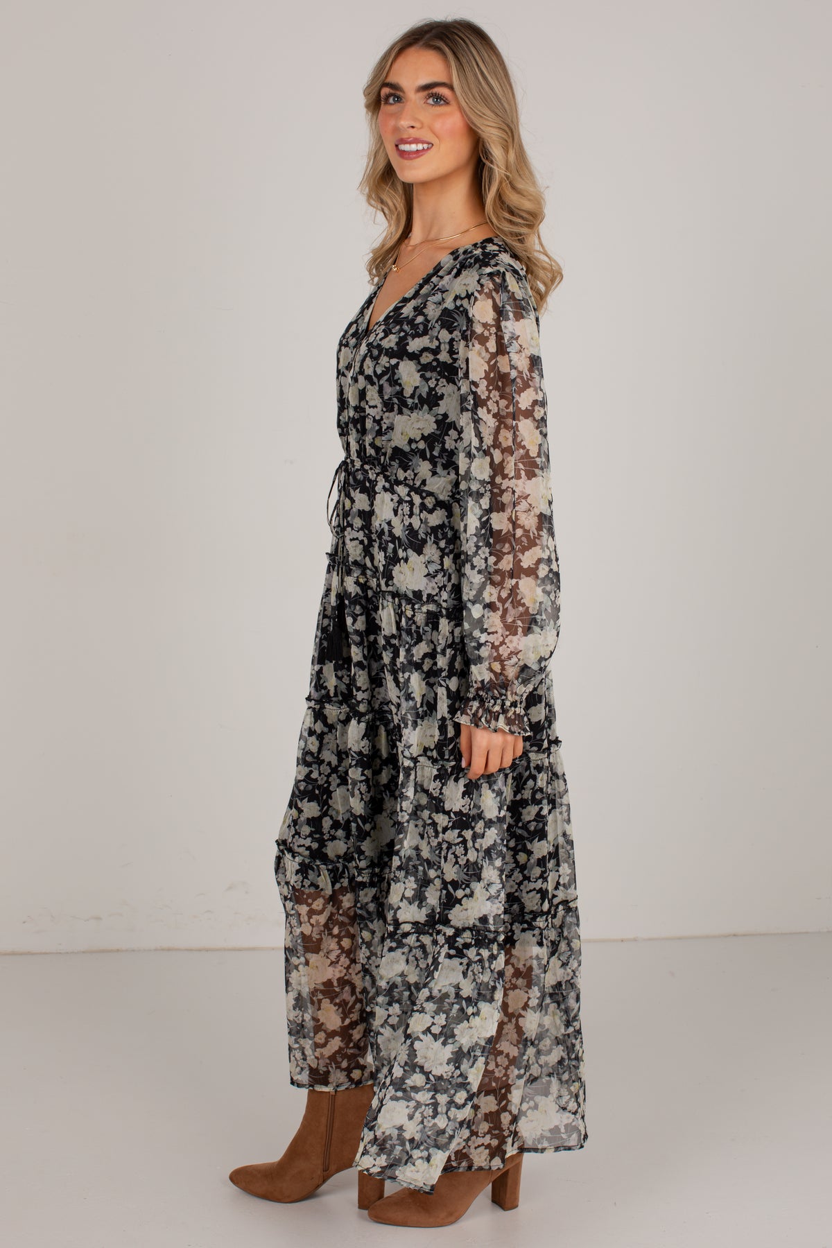 A woman modeling a long-sleeve maxi dress with a black, white, and gray floral pattern and sheer mesh panels, paired with brown ankle boots, against a plain light gray studio background.