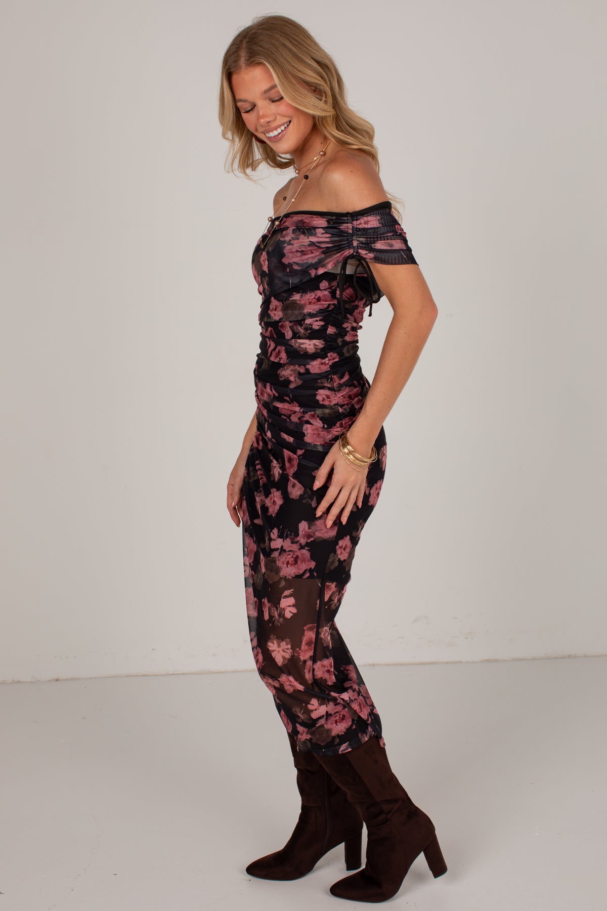 A woman modeling an off-the-shoulder black jumpsuit with a pink floral pattern, paired with dark brown heeled boots, against a plain light gray studio background.