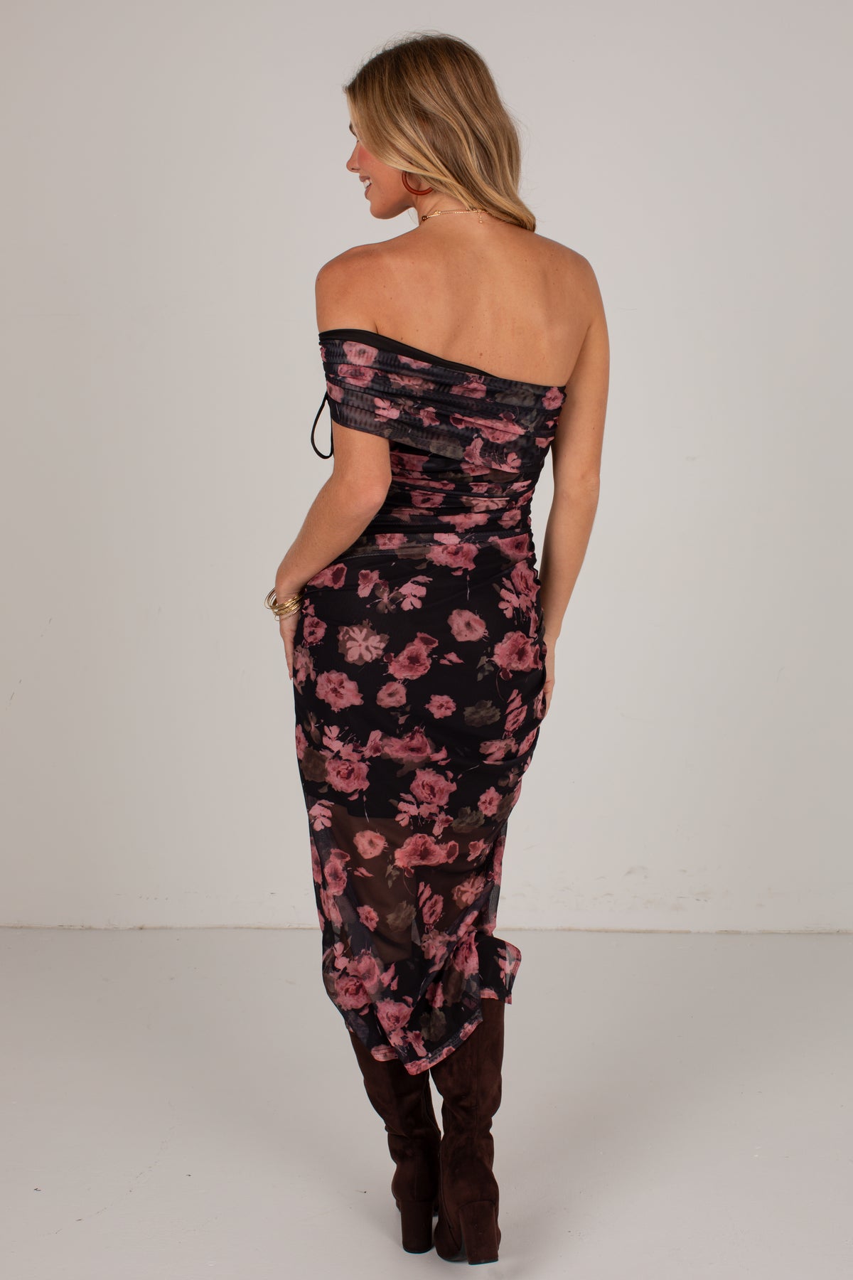 A woman shown from the back wearing a black off-the-shoulder midi dress with a pink floral print and dark brown knee-high boots, set against a plain light gray studio background.