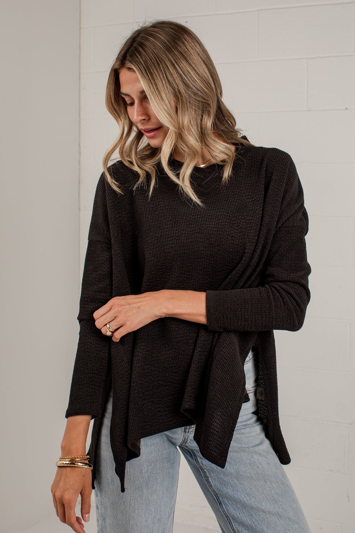 A woman wearing a dark brown long-sleeve knit top with a draped, asymmetrical hem, paired with light blue jeans, standing against a white brick wall.