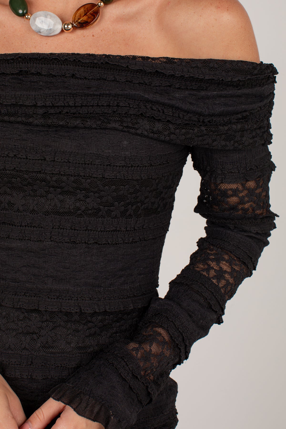 Close-up of a model wearing a black off-the-shoulder long-sleeve top with textured horizontal ruffles and sheer lace cuffs, paired with a chunky multi-colored statement necklace.