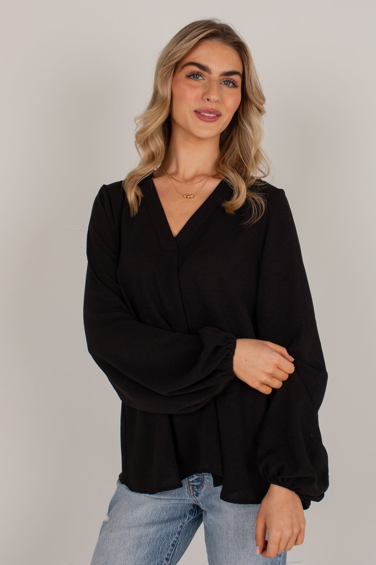 A woman modeling a black long-sleeve wrap-style blouse with a V-neck and gathered sleeves, paired with light blue jeans, against a plain light gray studio background.