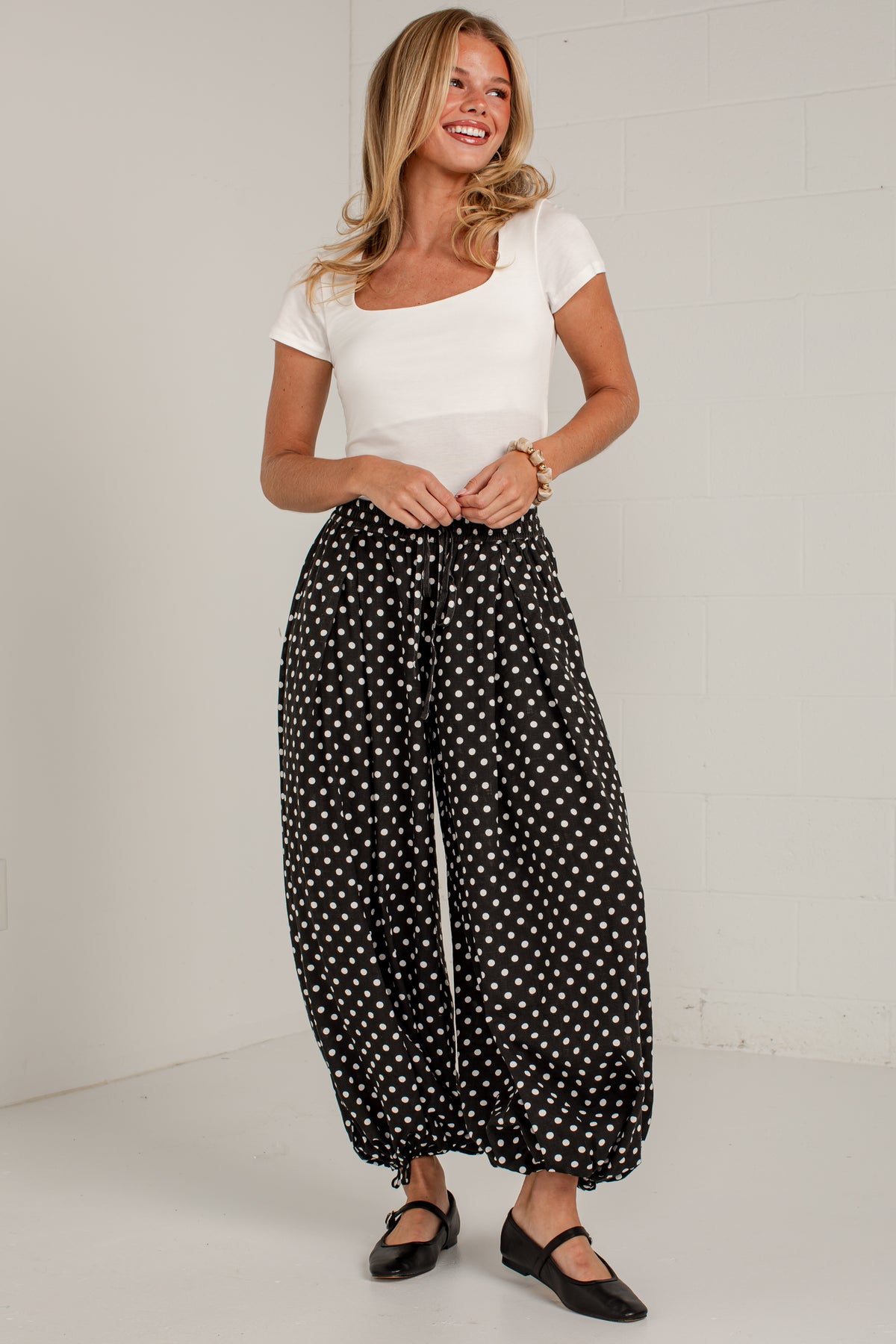 A woman modeling black wide-leg pants with a white polka dot pattern, paired with a white scoop-neck t-shirt and black Mary Jane shoes, standing against a white brick wall.