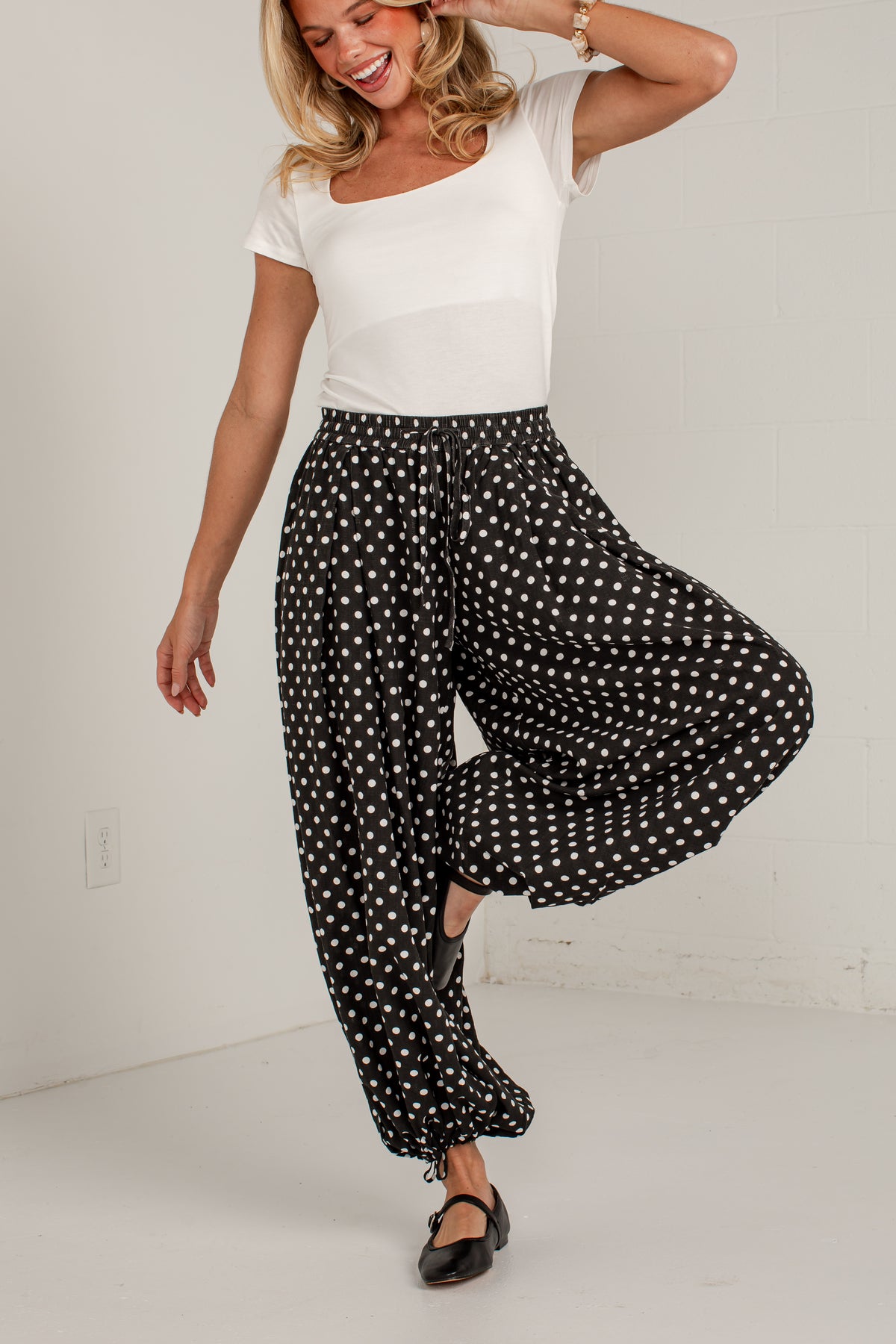 A woman modeling black wide-leg pants with white polka dots, paired with a white scoop-neck top and black flats, posing in a bright white room.