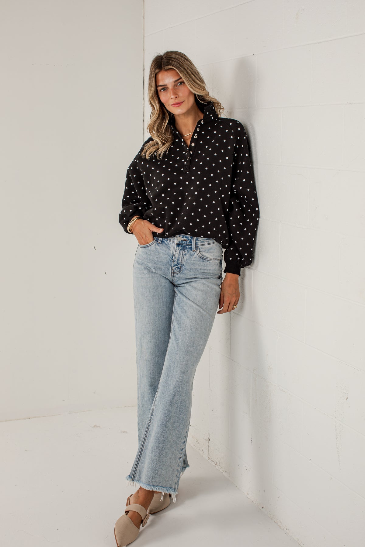 A woman stands in a white studio corner wearing a black long-sleeve blouse with white polka dots, light-wash flare jeans, and beige flat sandals.