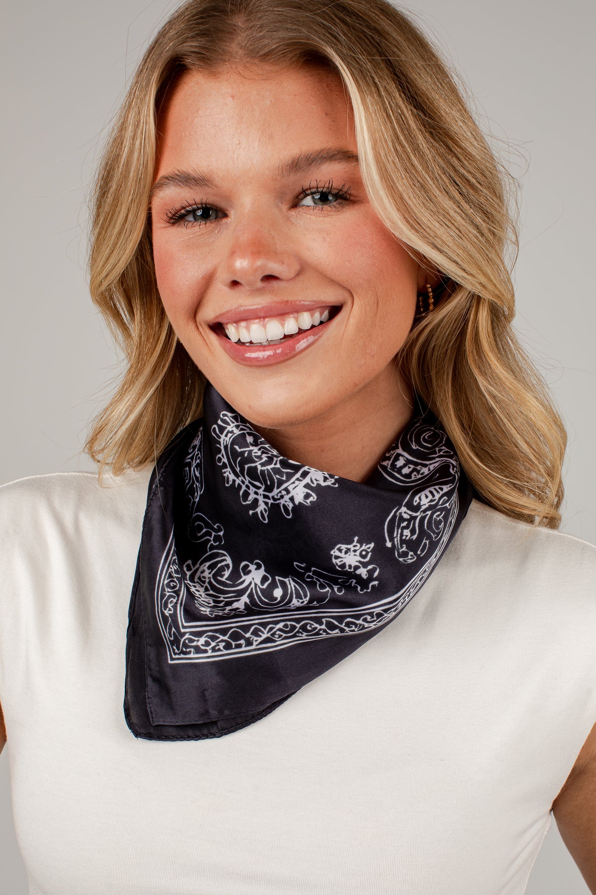 A smiling blonde woman wearing a black bandana with a white paisley pattern around her neck, paired with a white top, against a neutral gray background.