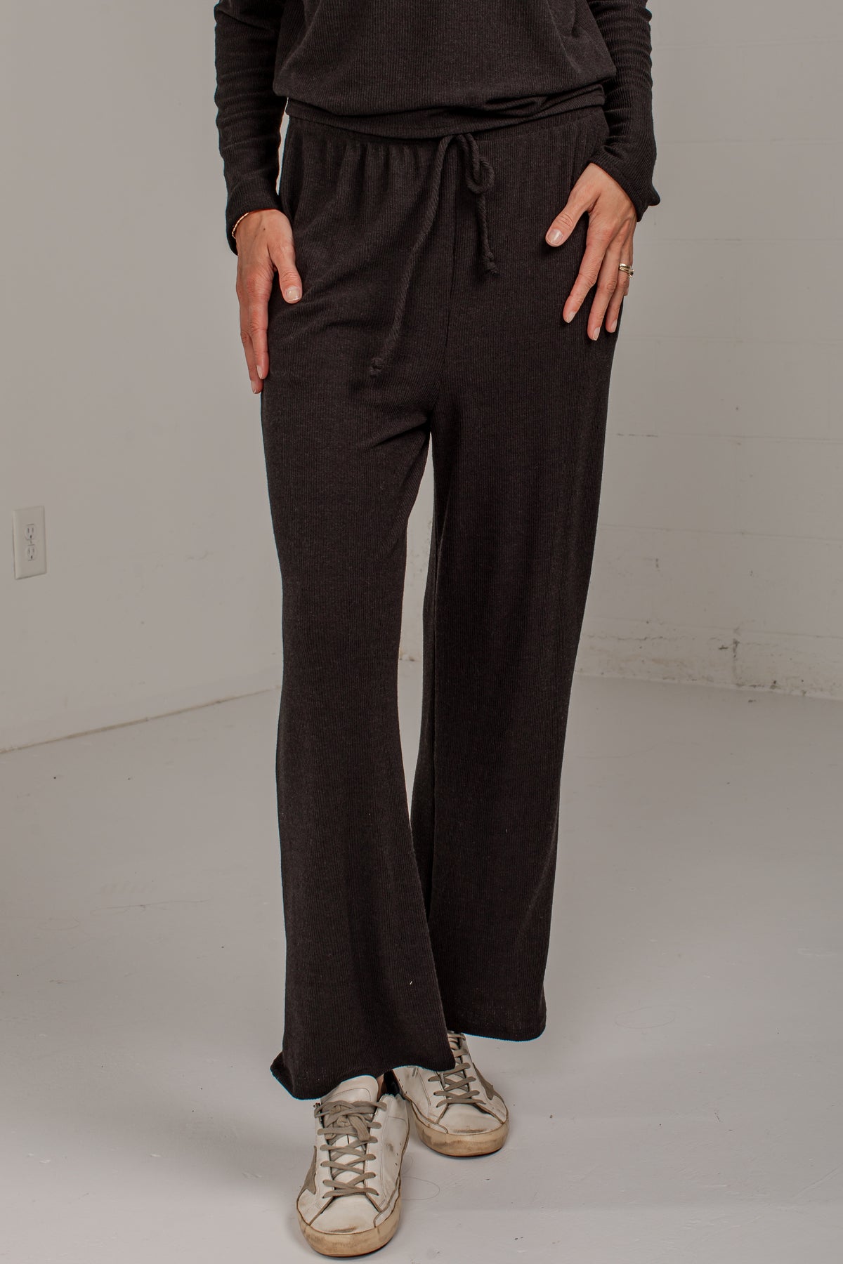 A person wearing dark brown wide-leg drawstring pants with a matching long-sleeve top and distressed white sneakers, standing in a plain studio setting.