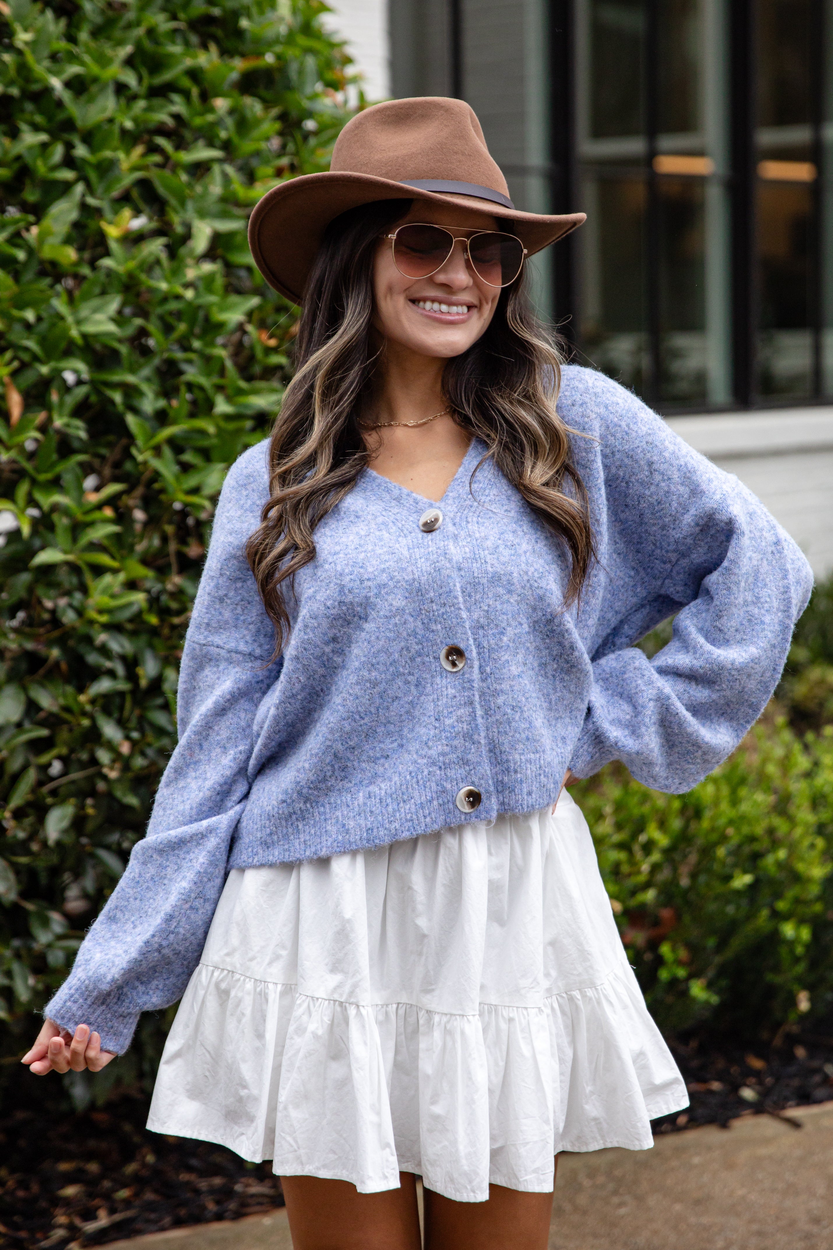 Woman wearing a blue cardigan, white skirt, and brown hat outdoors.