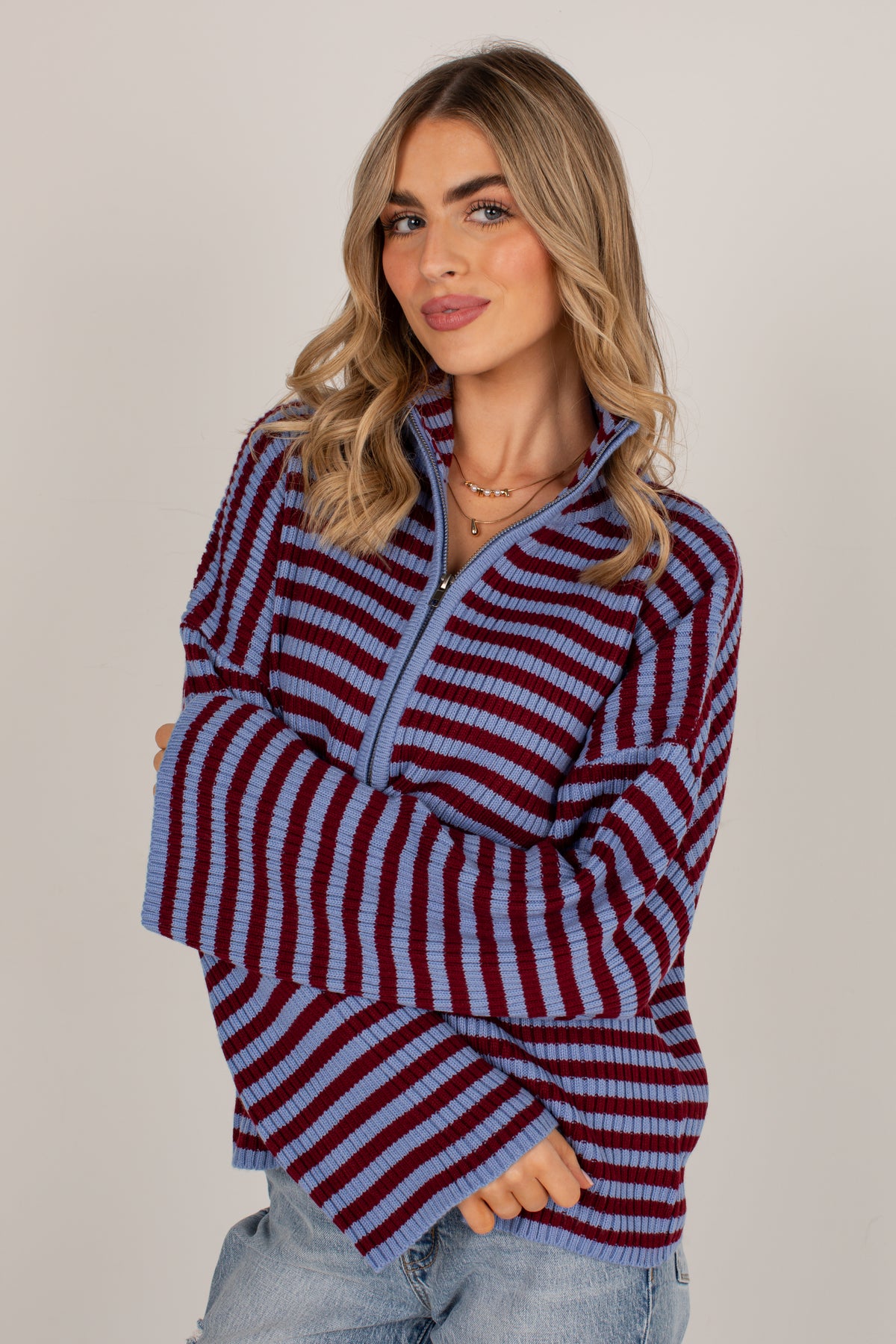 A woman wearing a long-sleeve quarter-zip knit sweater with horizontal burgundy and light blue stripes, paired with light-wash jeans, against a solid light gray studio background.