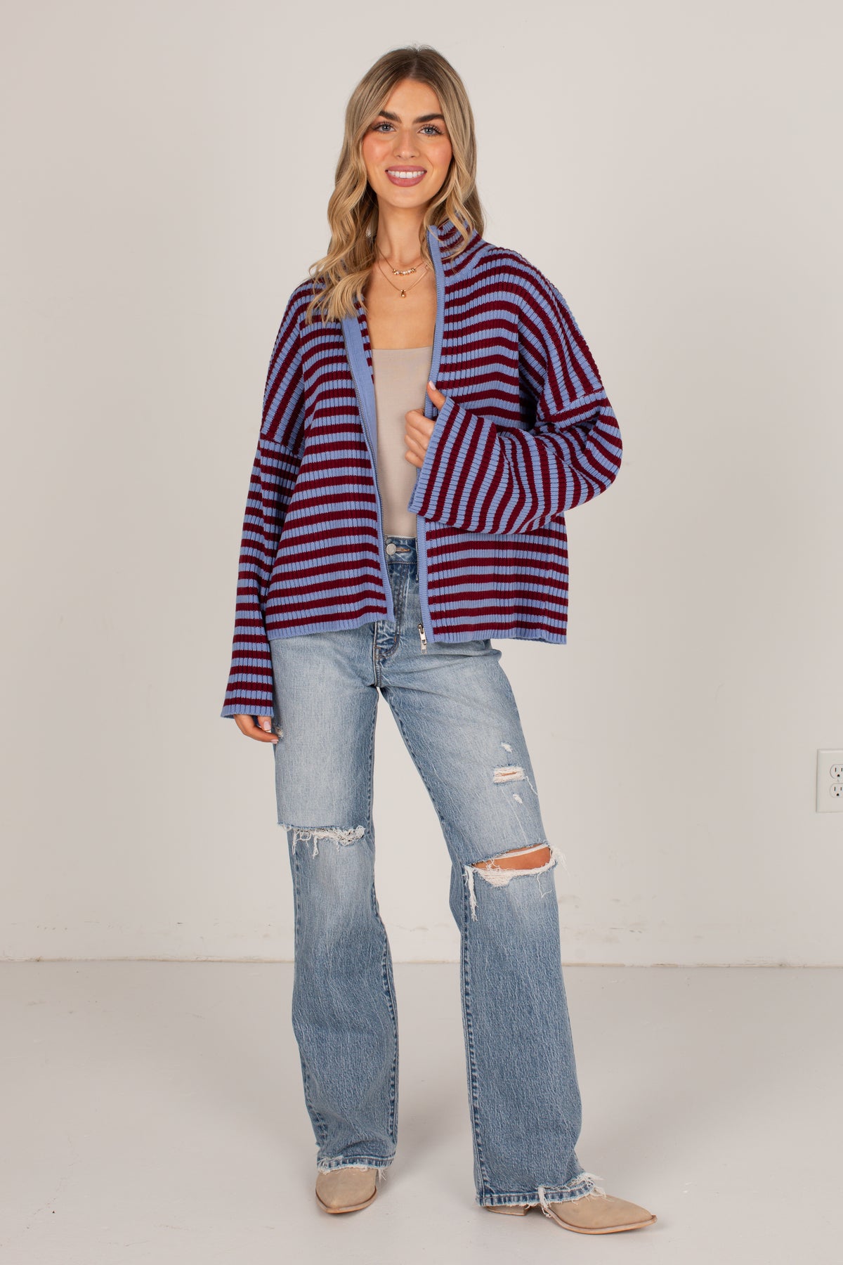 A woman modeling a striped cardigan with red and navy horizontal stripes, worn over a beige tank top and paired with light-wash distressed jeans and beige boots, against a plain off-white studio background.