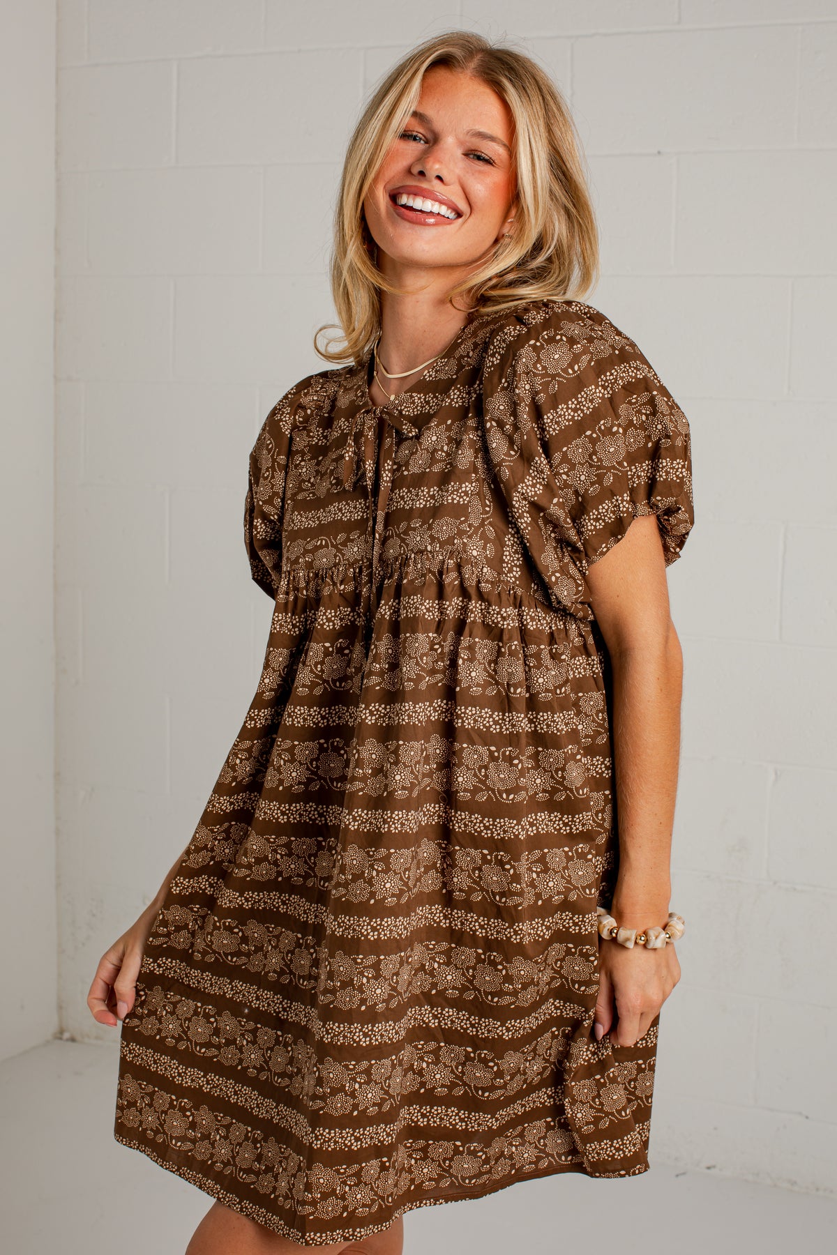 A smiling blonde woman wearing a brown and beige patterned short-sleeve dress, standing against a white brick wall.