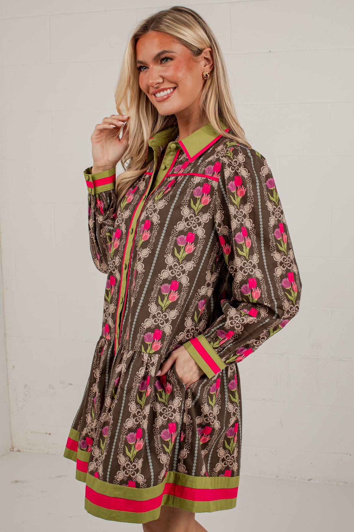 A woman modeling a brown long-sleeve shirt dress with a pink and green floral pattern, featuring lime green and hot pink striped cuffs and hem, set against a white brick wall.