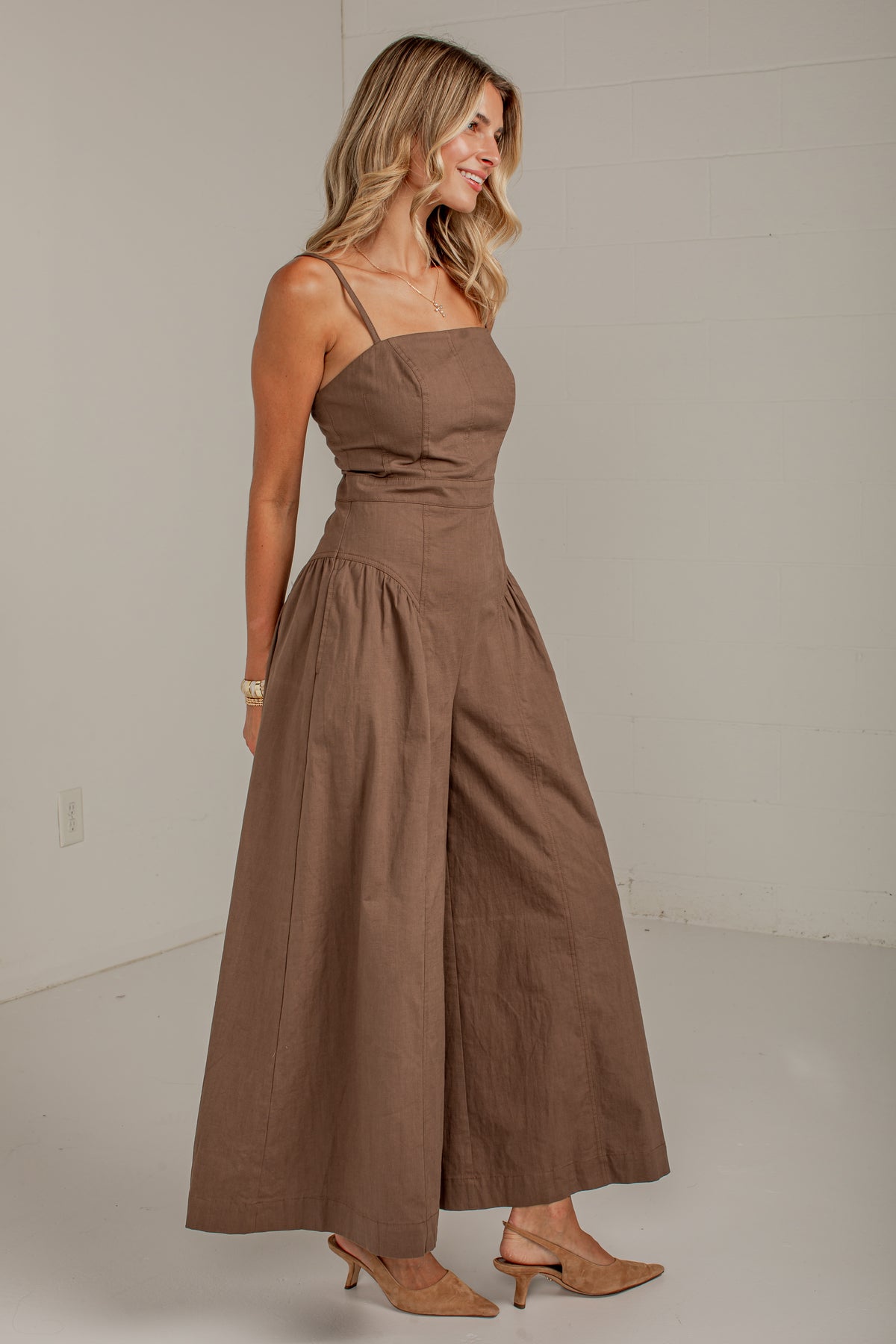 A woman modeling a brown sleeveless maxi dress with thin straps, a fitted bodice, and wide legs, standing in a plain indoor studio setting.