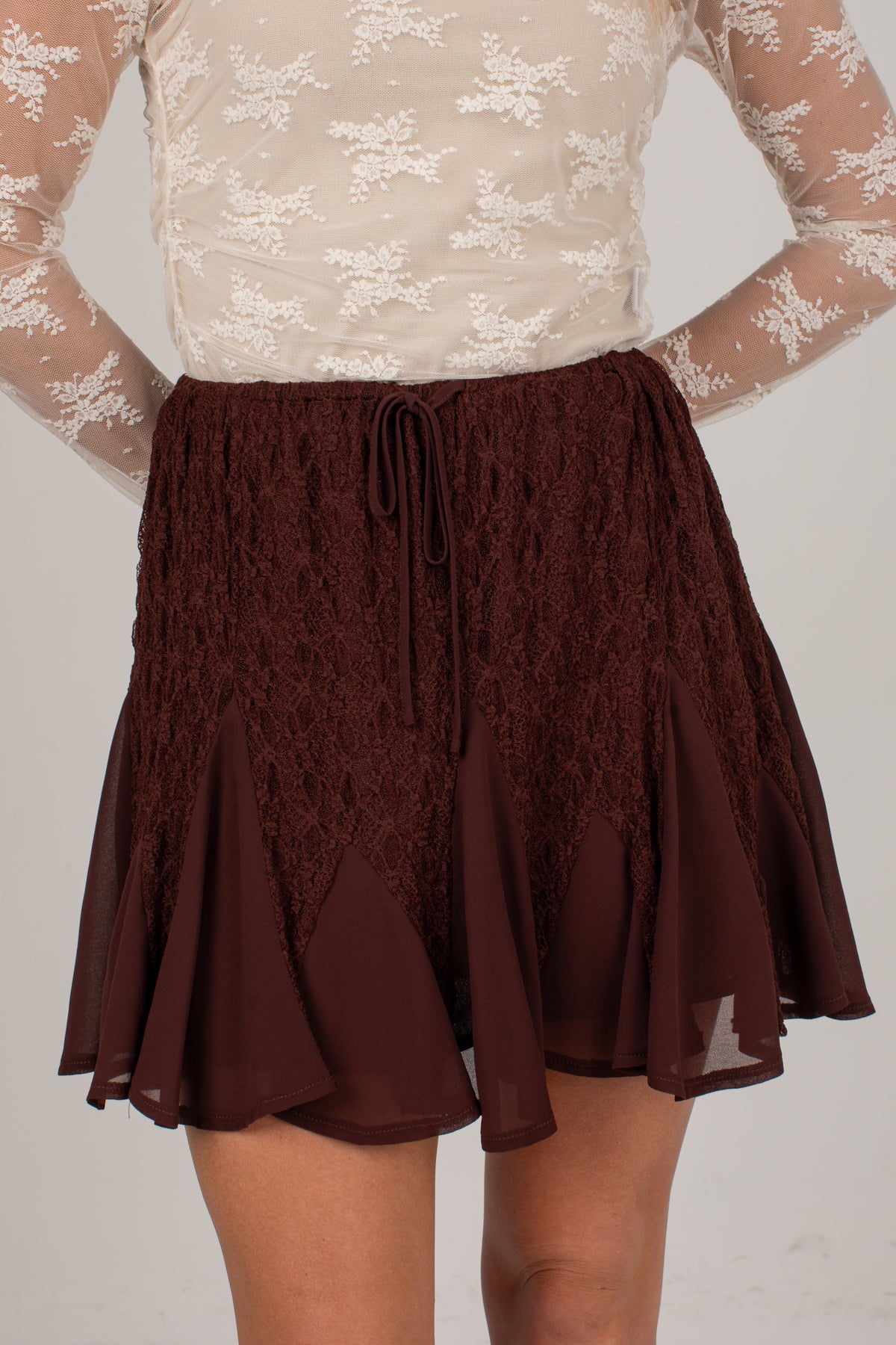 A close-up of a model wearing a dark brown lace and mesh mini skirt with a drawstring waist, paired with a beige lace top.