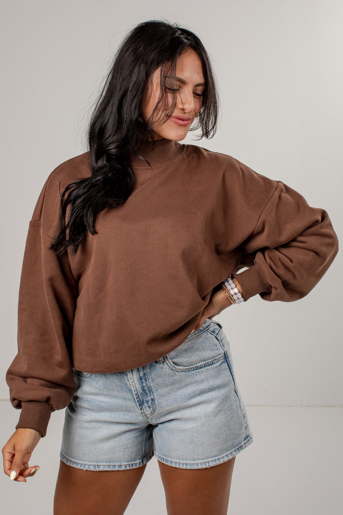 A woman wearing a brown long-sleeve mock neck sweatshirt and light-wash denim shorts, standing against a neutral light gray studio background.