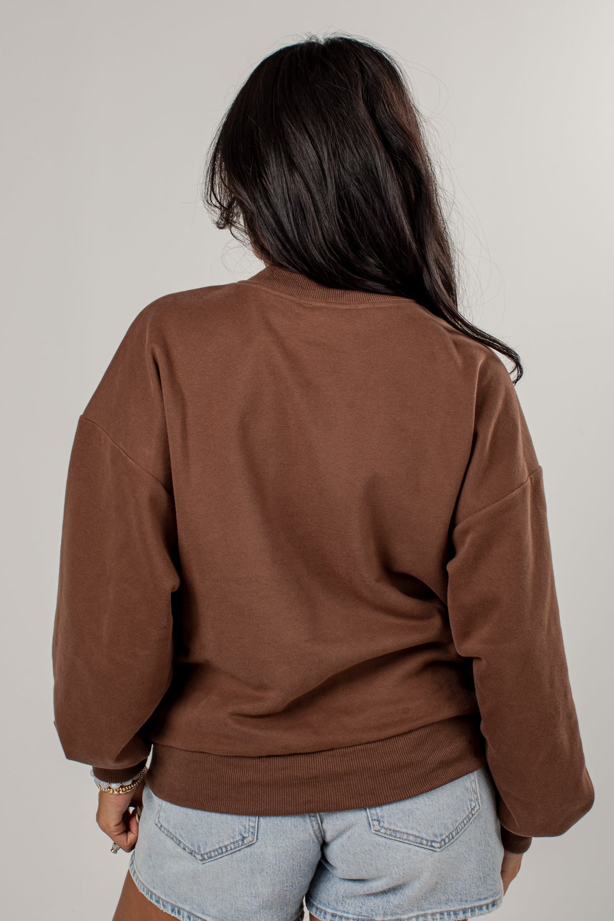 A woman viewed from behind wearing a brown crewneck sweatshirt and light blue denim shorts against a plain light gray background.