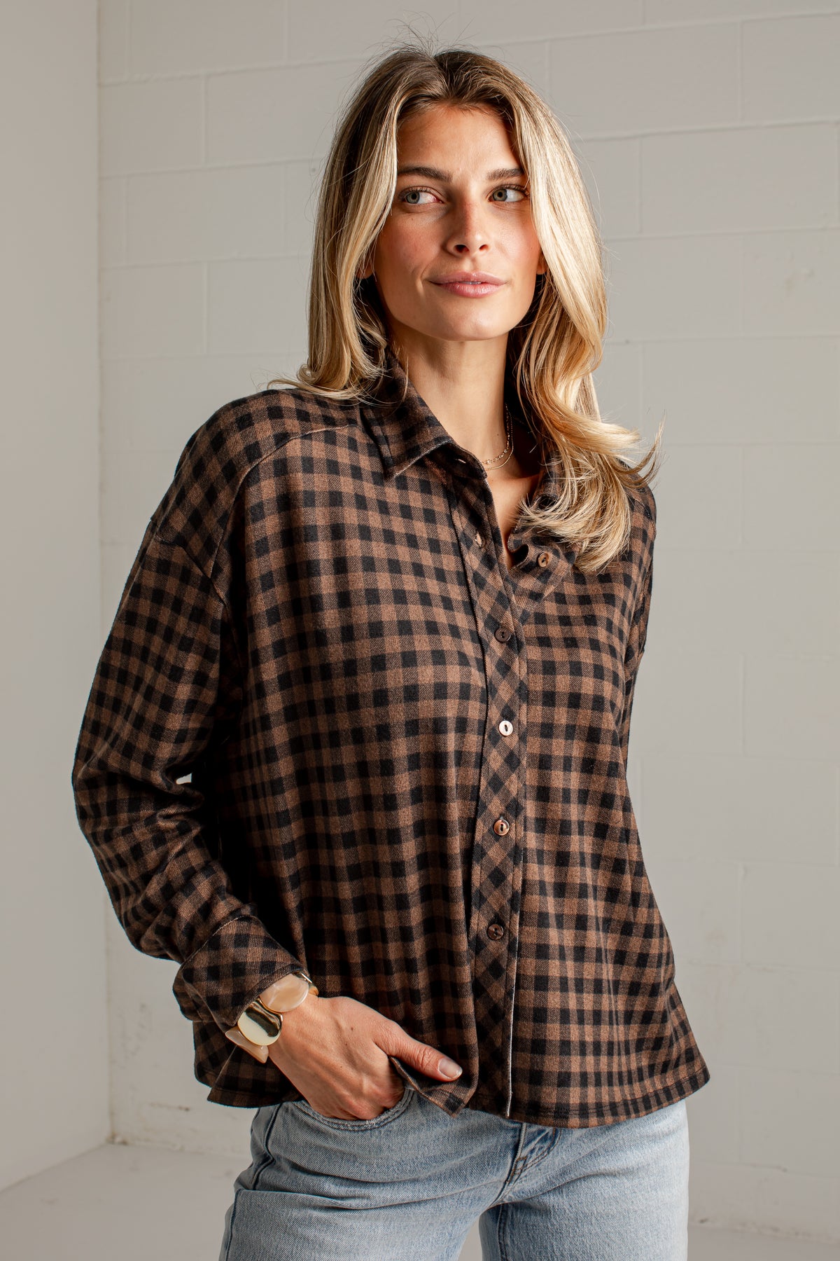 A woman wearing a brown and black checkered button-down flannel shirt with light blue jeans, standing against a white brick wall.