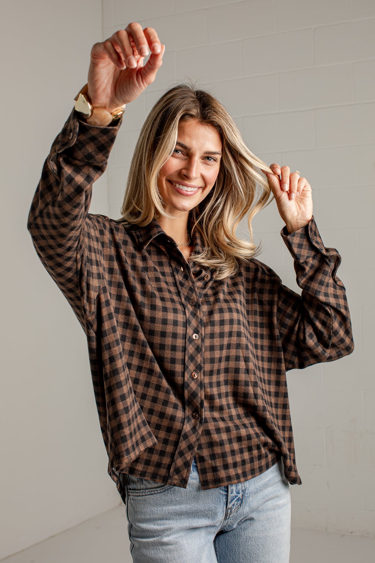 A woman wearing a brown and black checkered flannel button-up shirt and light blue jeans, posing with one arm raised against a light gray brick wall.