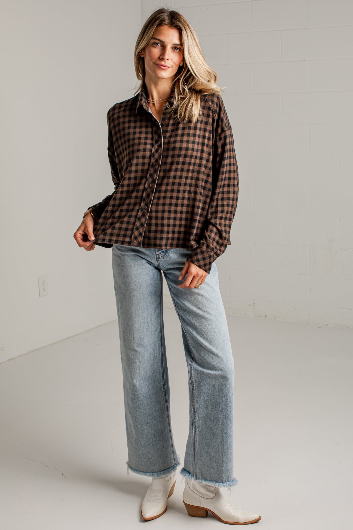 A woman modeling a brown and black checkered long-sleeve button-up shirt, paired with light blue wide-leg jeans and white cowboy boots, standing against a white studio wall.