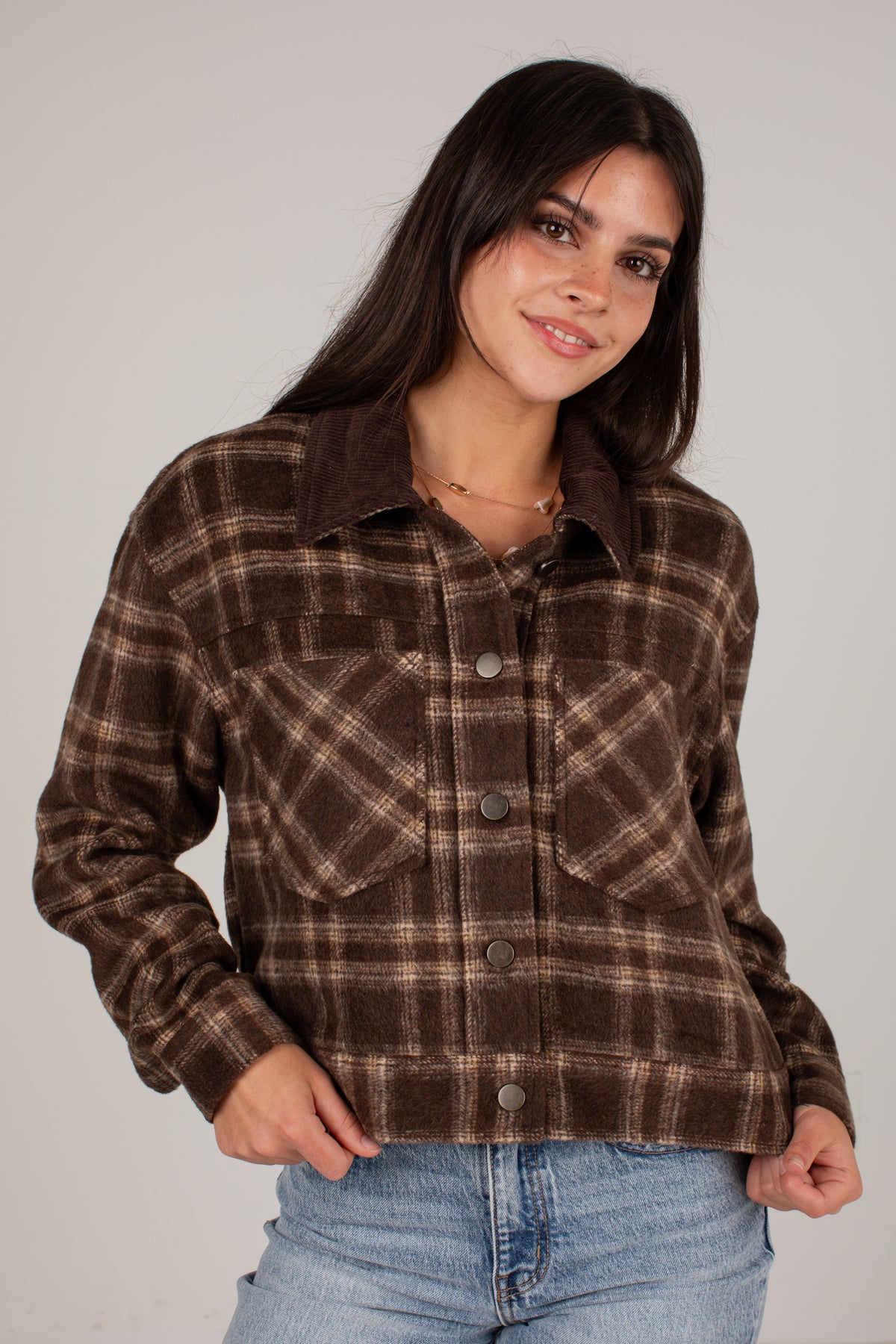 A woman wearing a brown and beige plaid button-up flannel shirt jacket with a dark brown corduroy collar, paired with light blue jeans, against a plain light gray background.