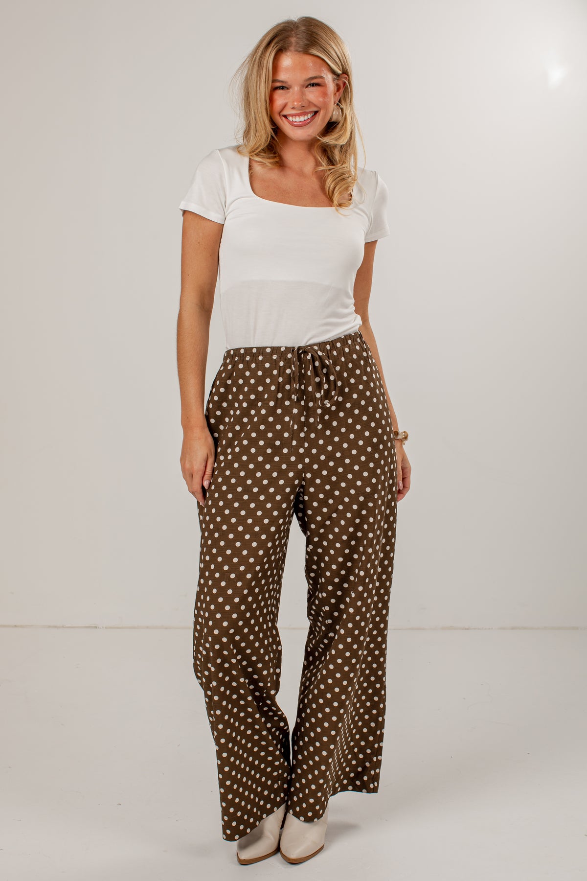 A woman modeling a white short-sleeve top and brown wide-leg pants with a white polka dot pattern, standing against a plain white studio background.