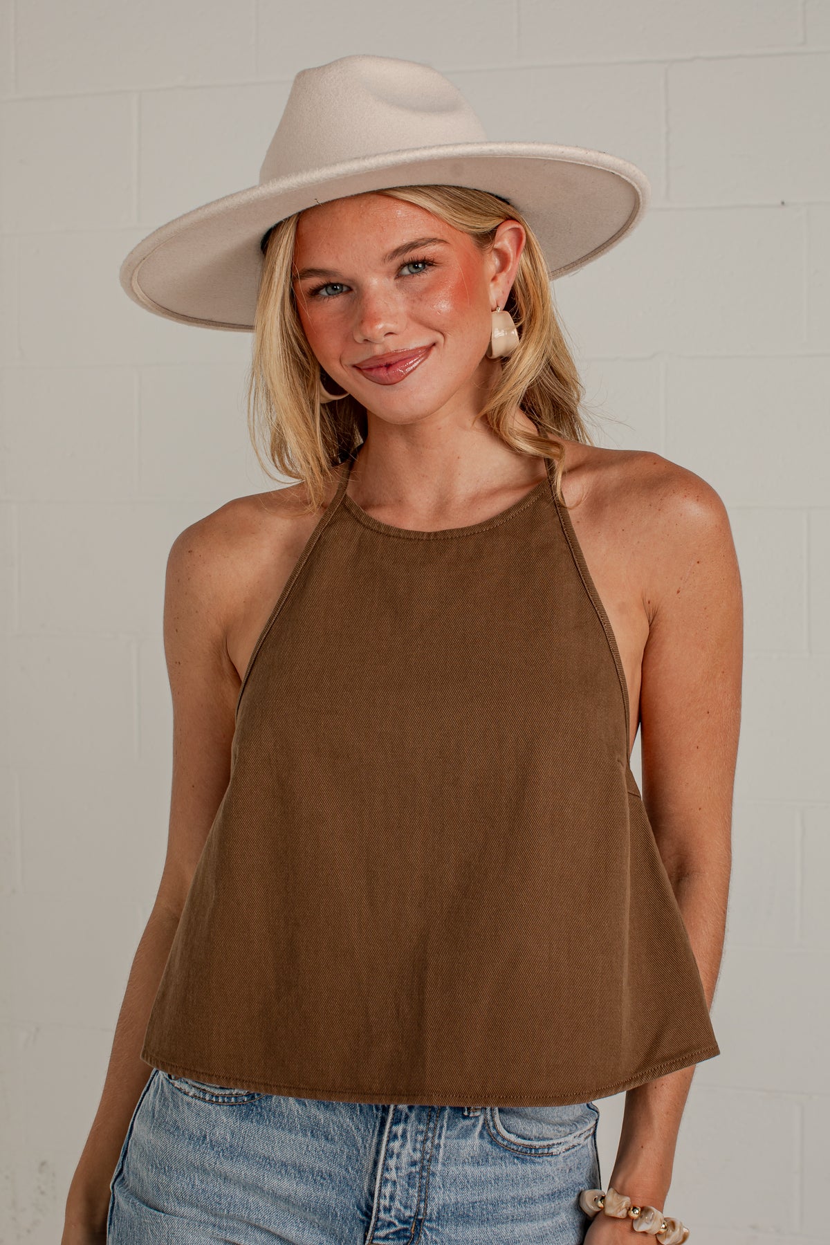 A woman wearing a brown halter crop top, a wide-brimmed cream hat, and light-wash blue jeans, standing against a white brick wall.