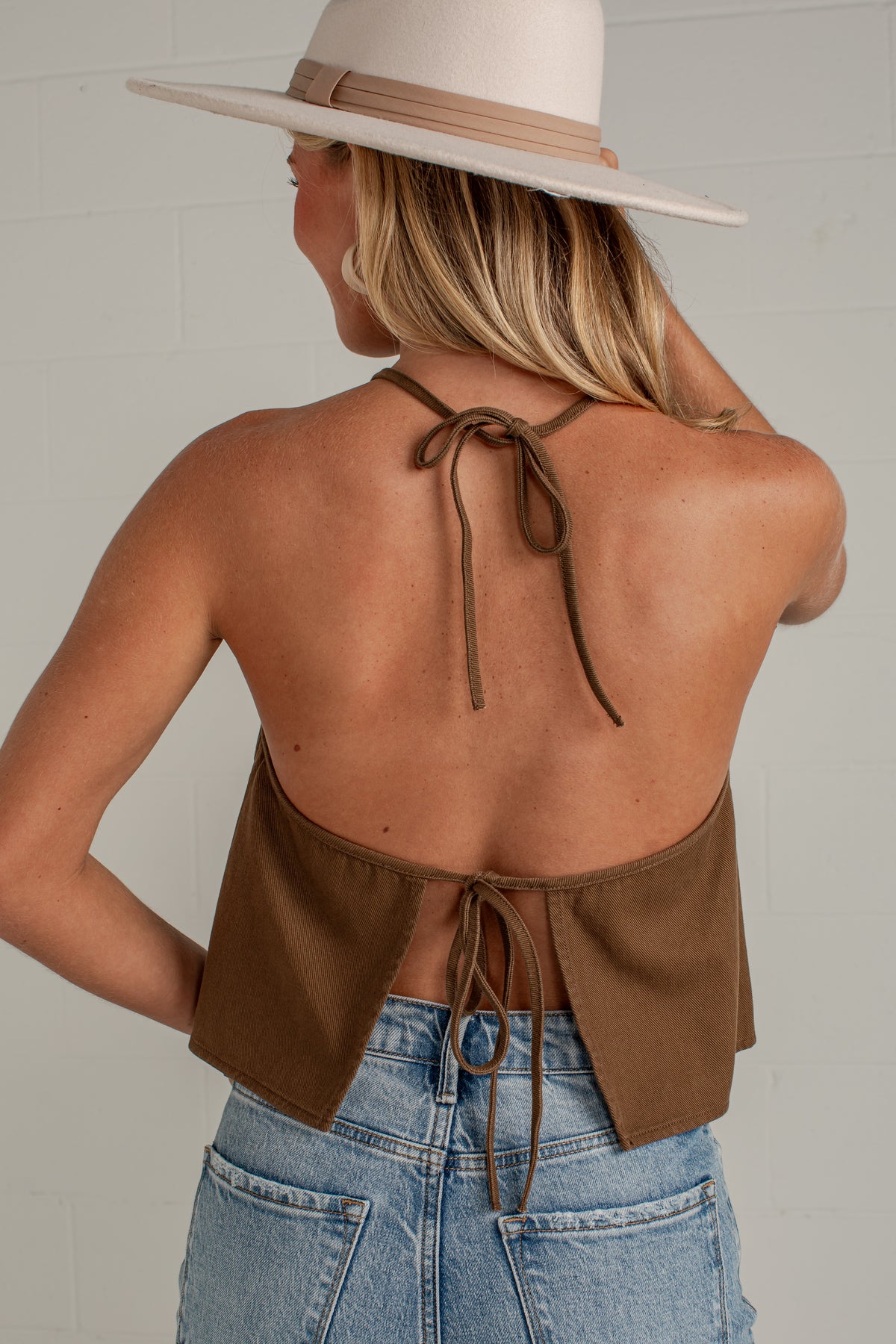 A woman shown from the back wearing a brown halter-style crop top with tie-back straps and light blue jeans, standing against a light gray wall.