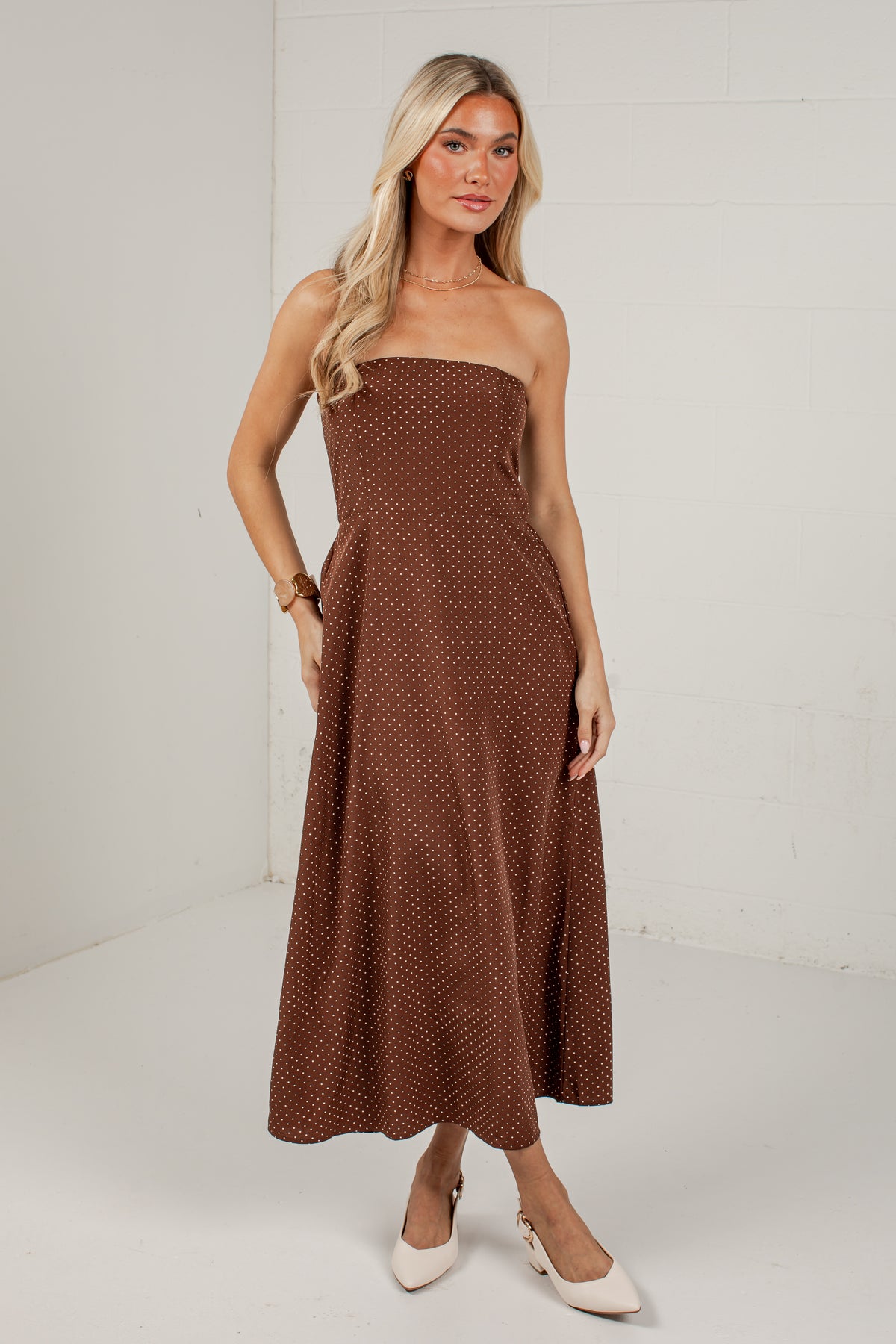 A woman modeling a strapless brown midi dress with a small white polka dot pattern, standing against a white brick wall background.
