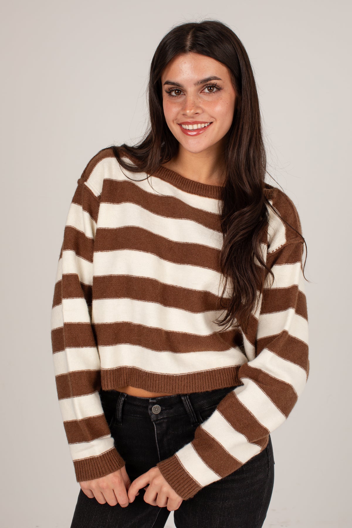 A woman modeling a brown and cream striped cropped knit sweater with black jeans, photographed against a plain light gray studio background.