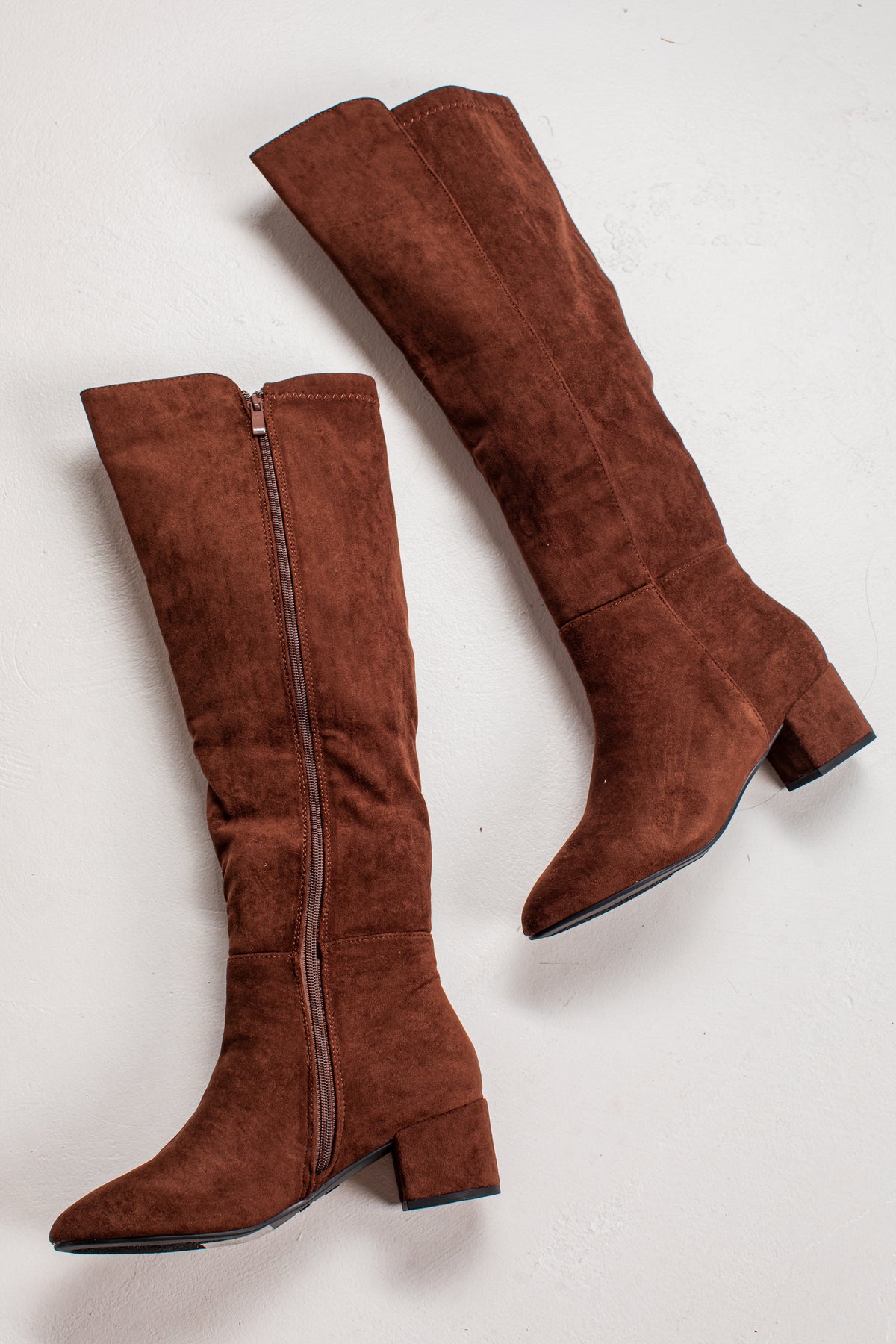 A pair of knee-high brown suede boots with side zippers and block heels, displayed on a light off-white background.