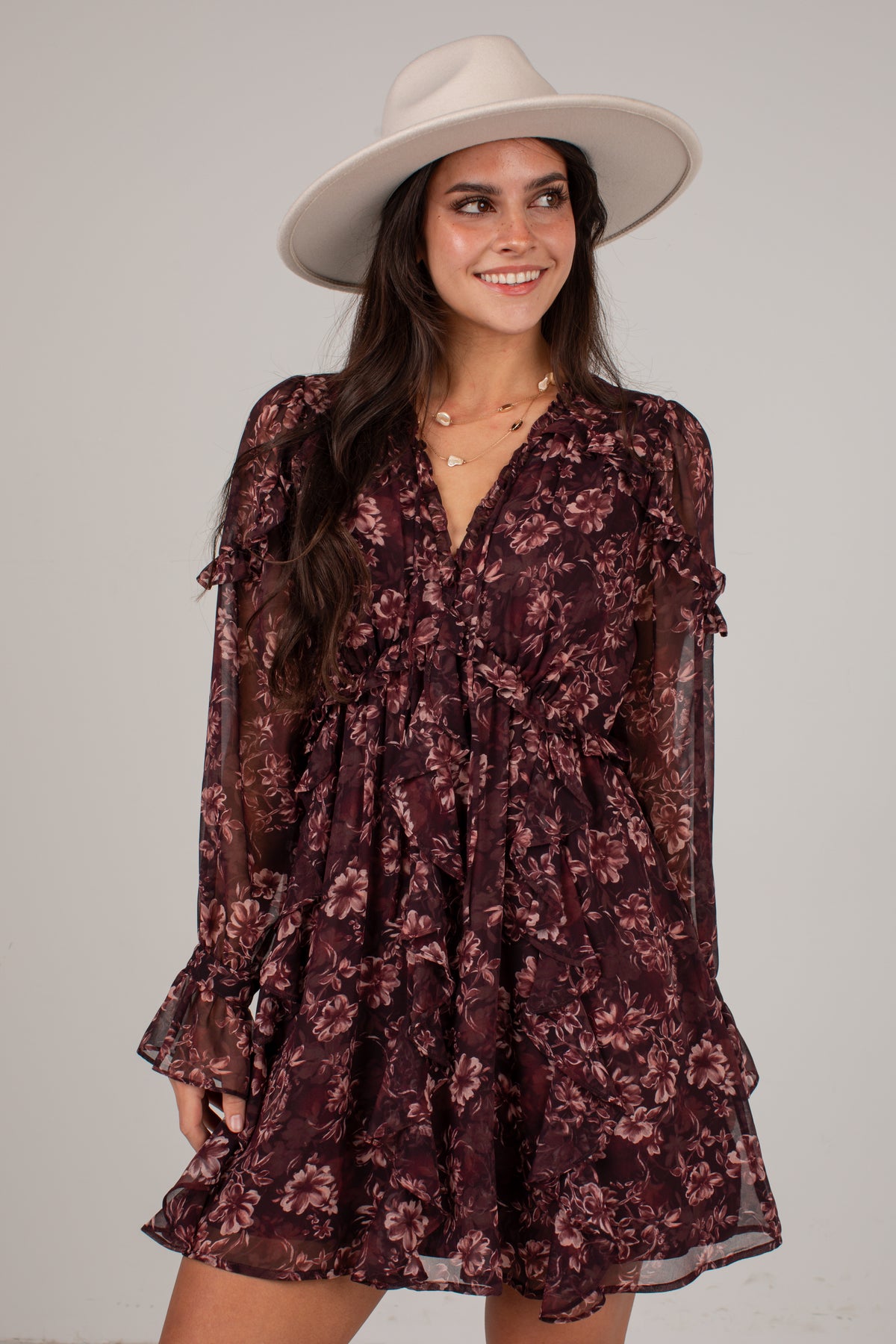 A woman wearing a dark burgundy floral mini dress with sheer long sleeves and a wide-brimmed cream hat, posing against a light gray studio background.