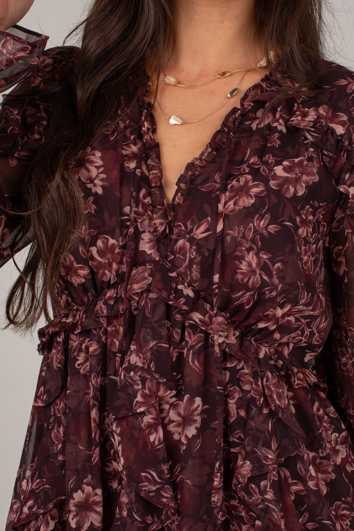 Close-up of a woman wearing a dark floral dress with a ruffled V-neckline and long sleeves, paired with layered gold necklaces.