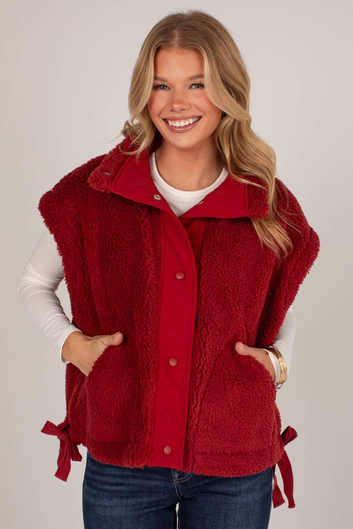 A smiling blonde woman wearing a red sherpa fleece button-front vest over a white long-sleeve shirt and blue jeans, photographed against a plain light gray studio background.