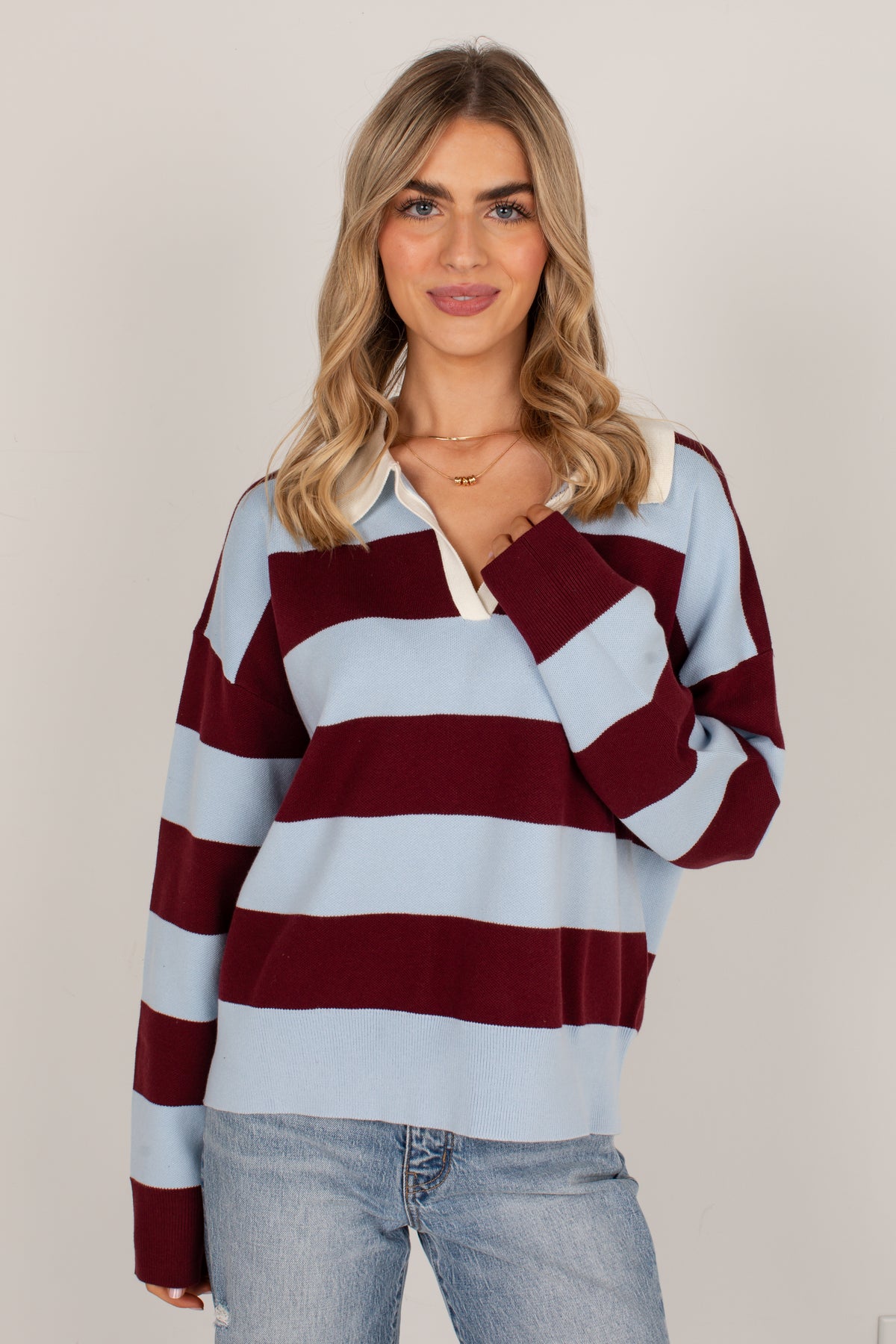 A woman wearing a light blue and burgundy striped rugby sweater with a white collar, paired with light-wash blue jeans, against a neutral beige background.