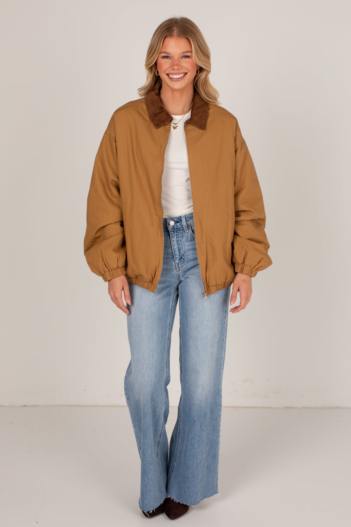A woman modeling a tan utility jacket with a brown corduroy collar, worn over a white top and light blue wide-leg jeans, against a plain off-white studio background.