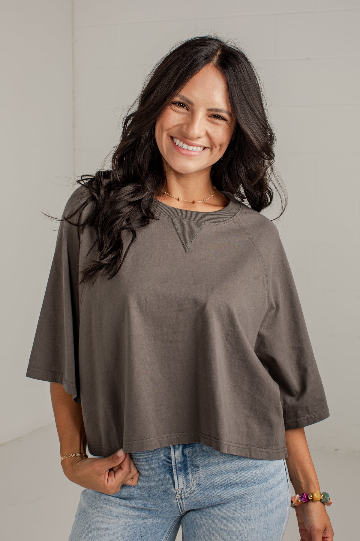 A woman with long dark hair smiling while wearing a loose-fitting, short-sleeve taupe-brown top and light-wash blue jeans against a light gray wall.